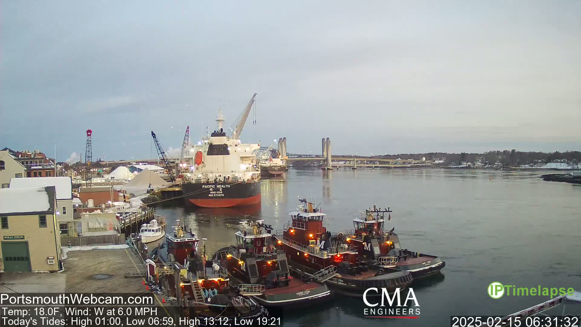 A large cargo ship and several tugboats are docked at a waterfront facility on a calm, cool day.