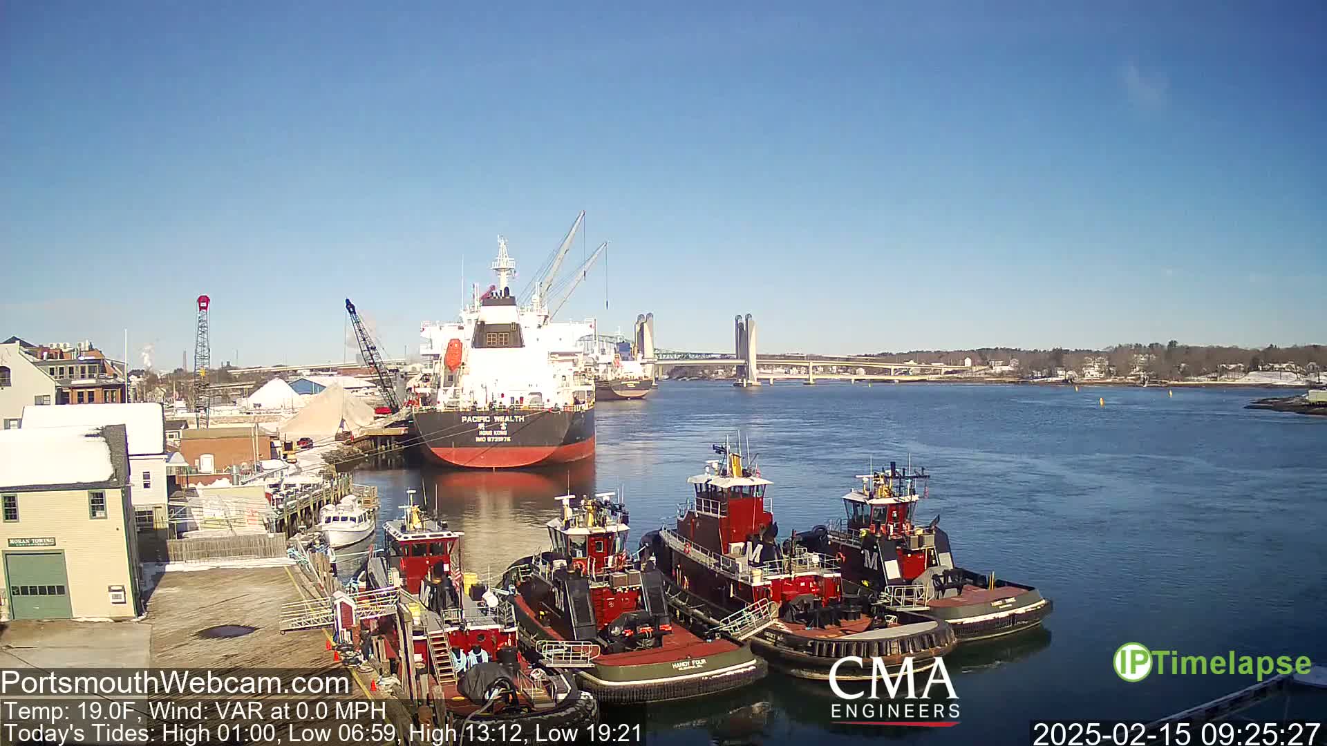 A large cargo ship is docked near several tugboats in a harbor under a clear blue sky.