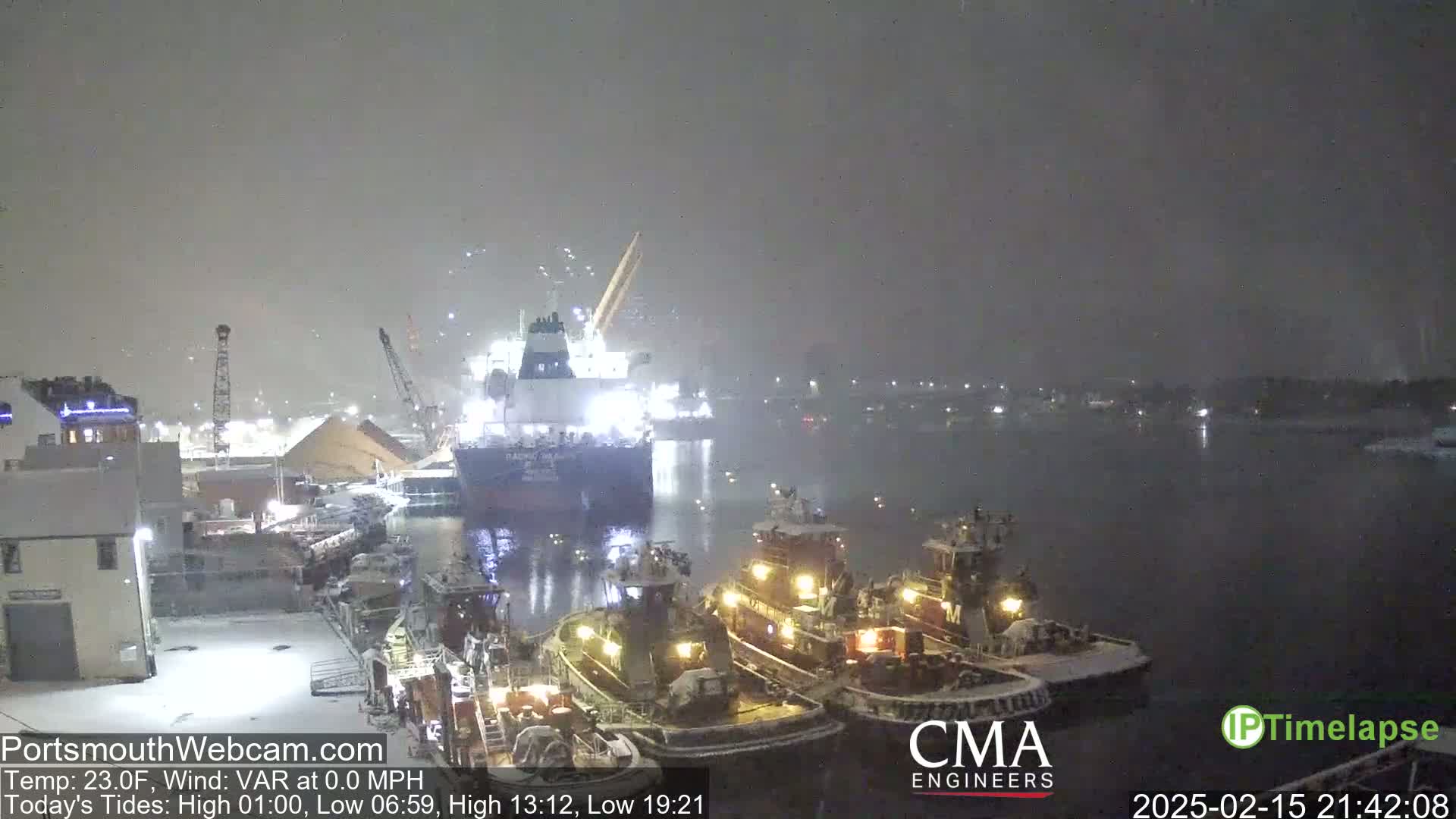 A large cargo ship and several tugboats are docked in a snowy harbor at night.