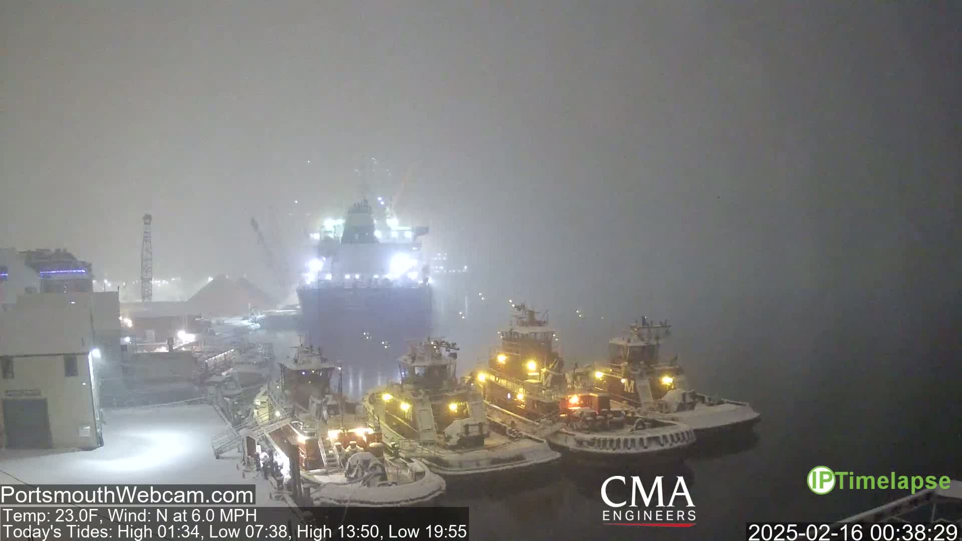 A large ship and several smaller tugboats are docked in a snowy harbor during a light snowstorm.