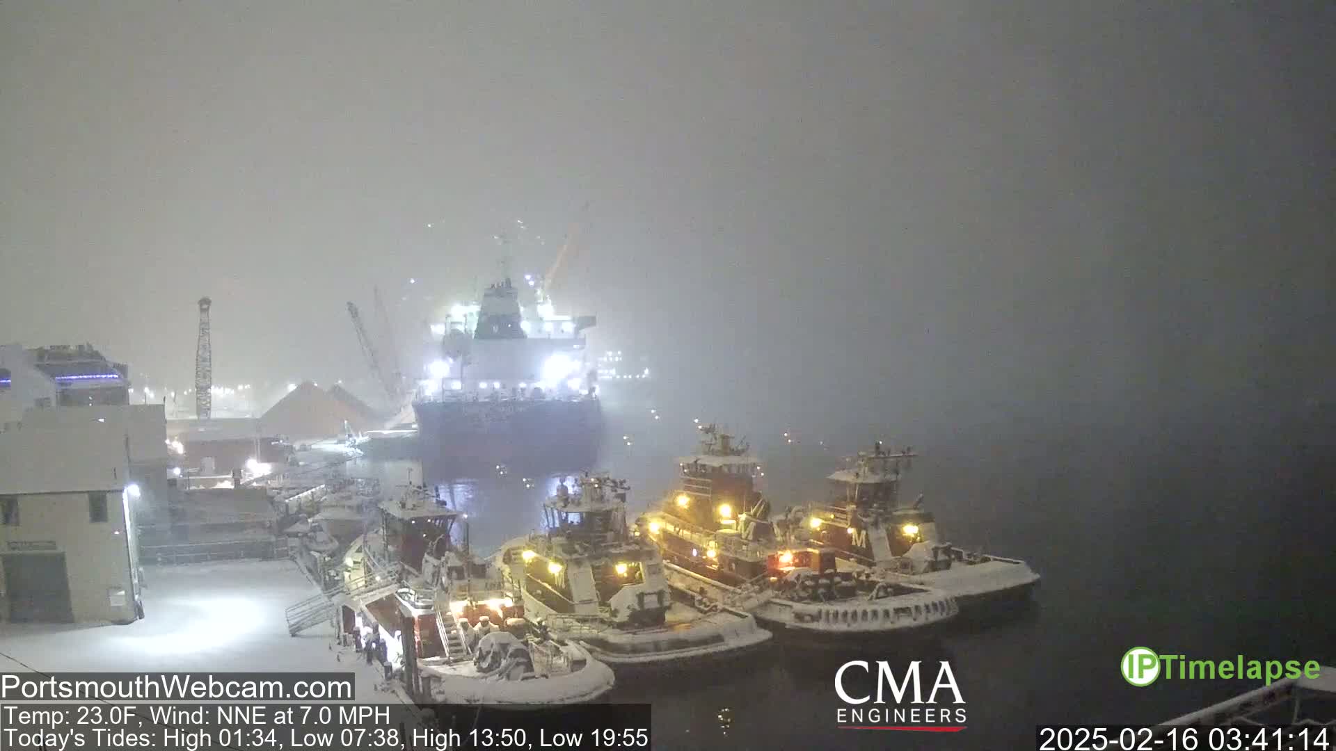A large cargo ship and three tugboats are docked in a snowy harbor during a heavy snowstorm.