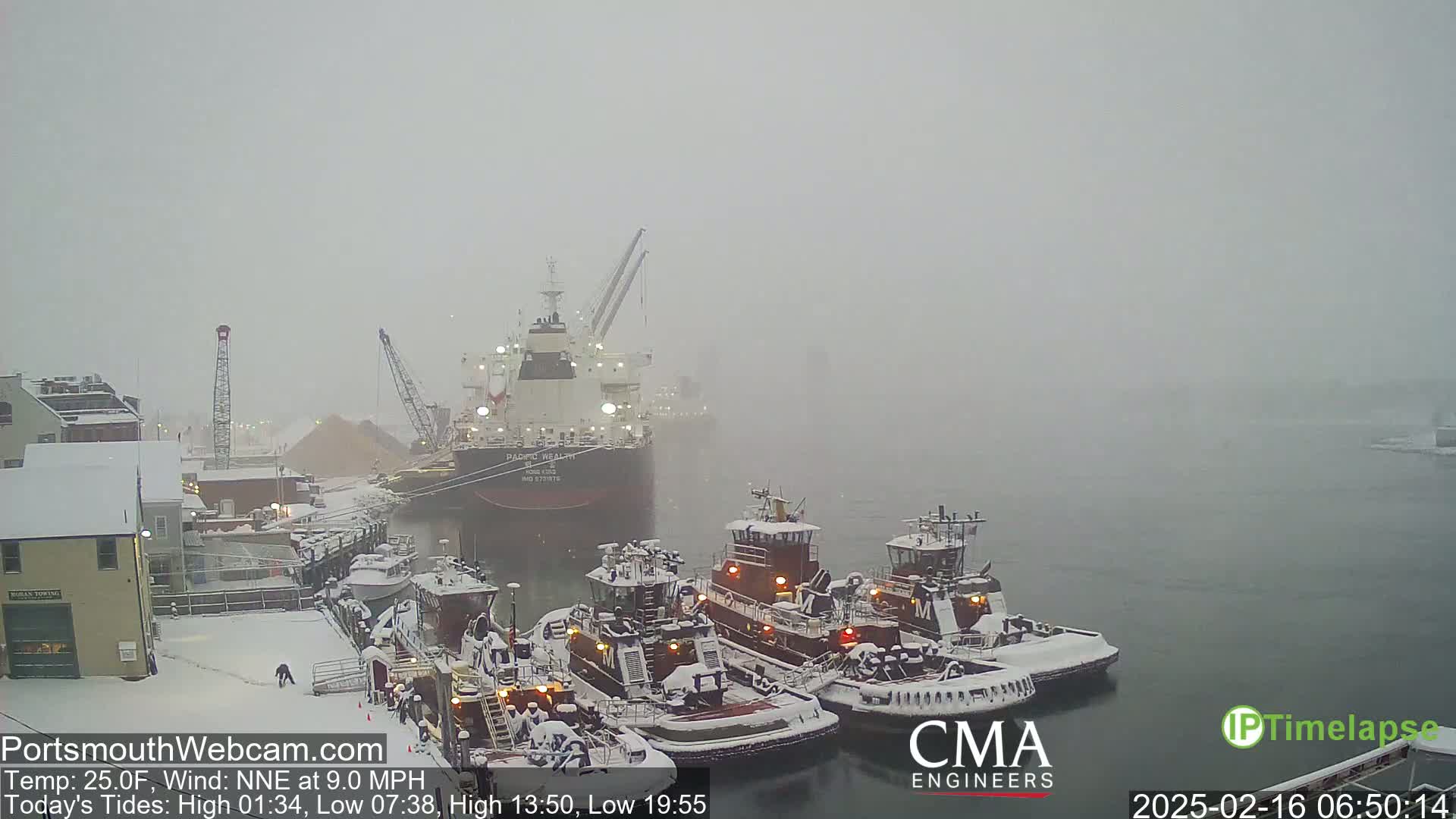 A large cargo ship and several tugboats are docked in a snowy harbor during a heavy snowstorm.