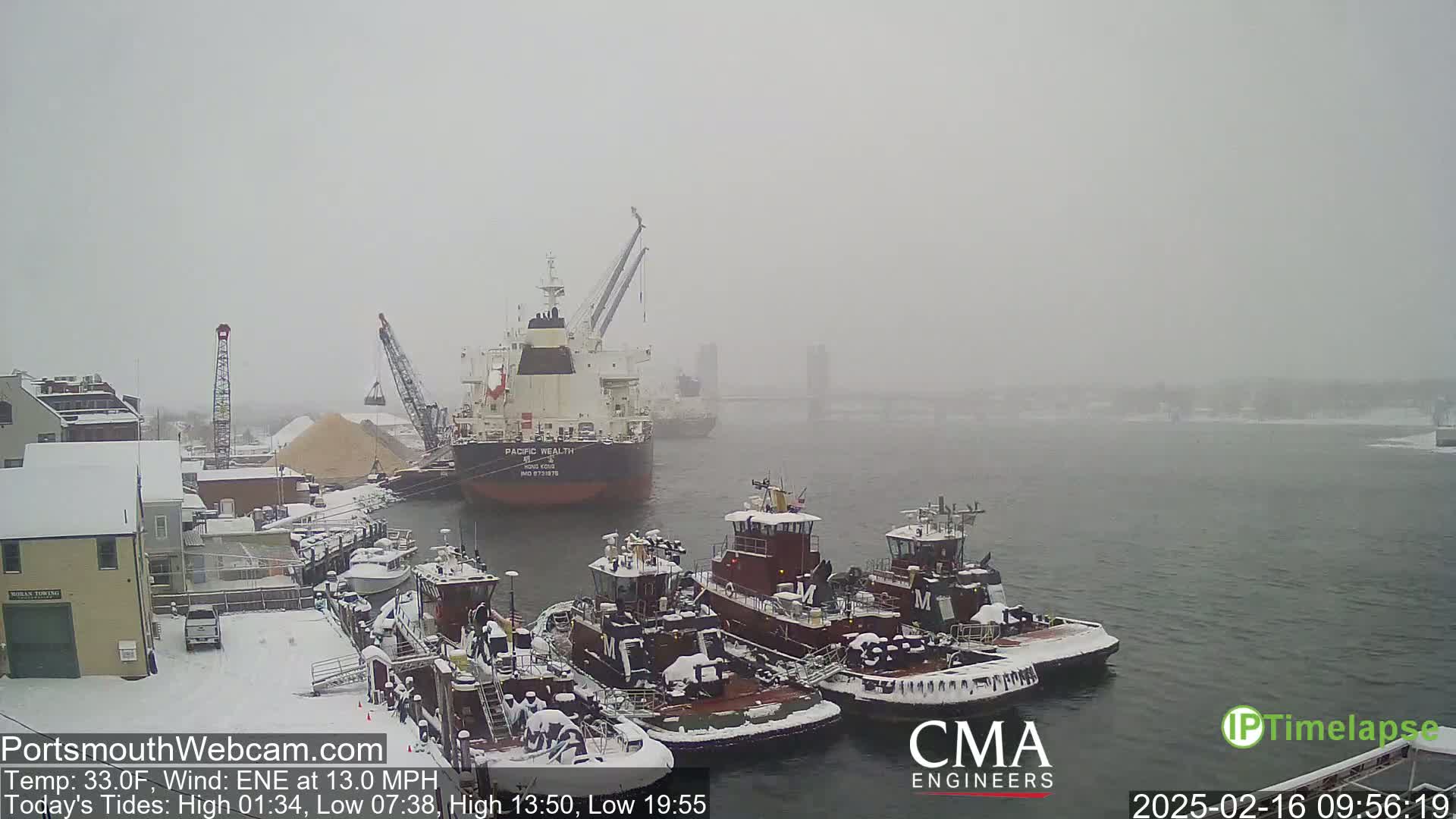 A large cargo ship and several tugboats are docked at a snow-covered harbor in snowy conditions.