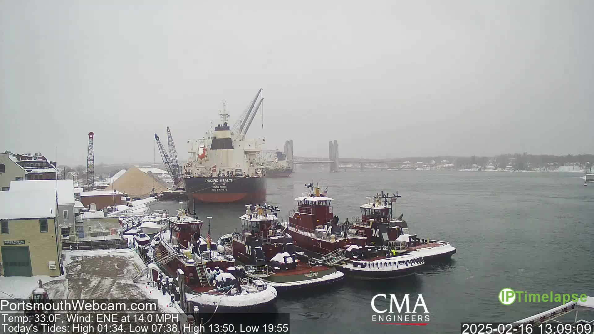 A snow-covered harbor scene shows several tugboats moored near a large cargo ship, with a bridge visible in the distance under a heavy, overcast sky.