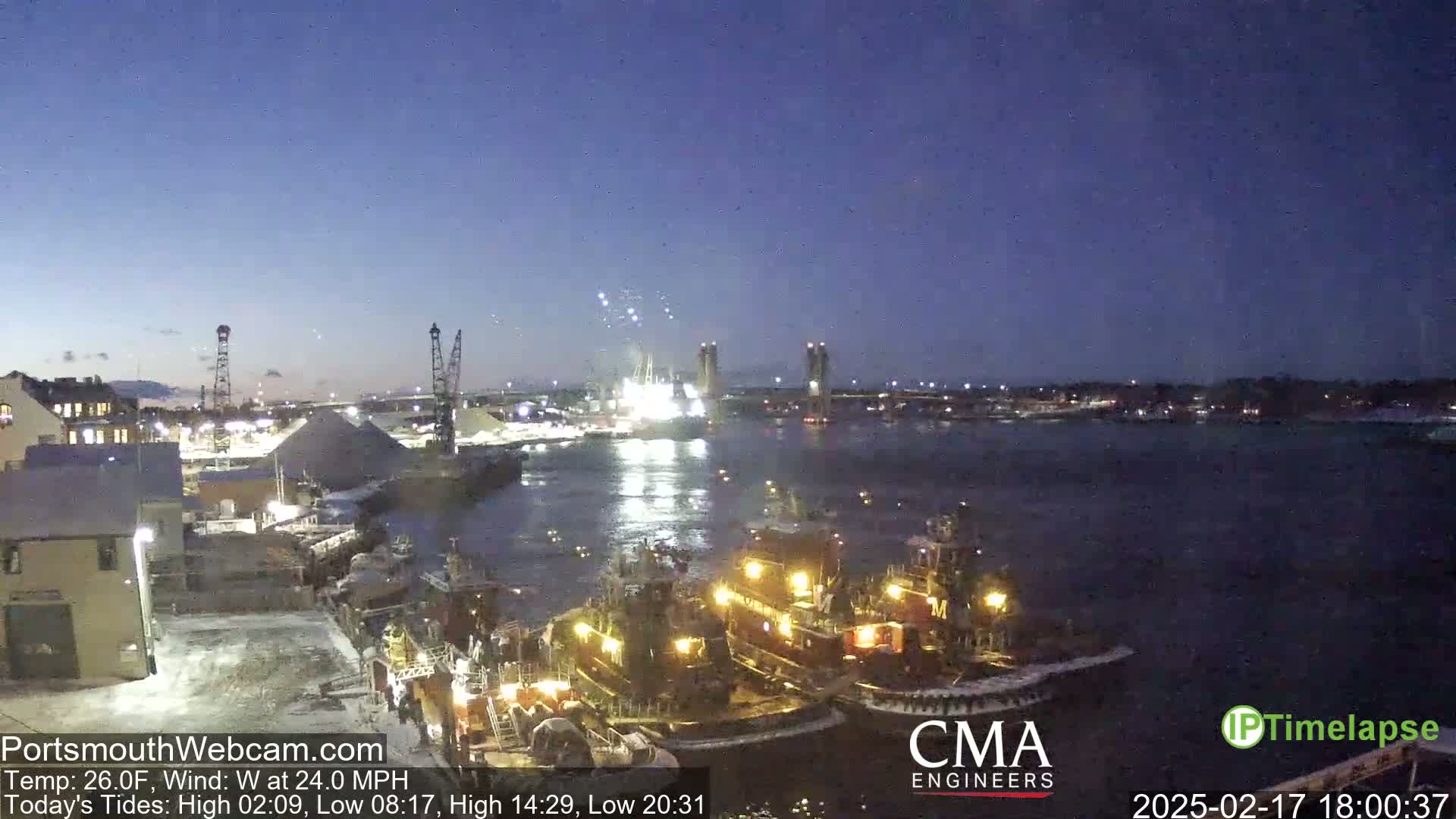 Several tugboats are docked at a harbor at twilight under a clear, dark blue sky, with a bridge and industrial structures visible in the background.
