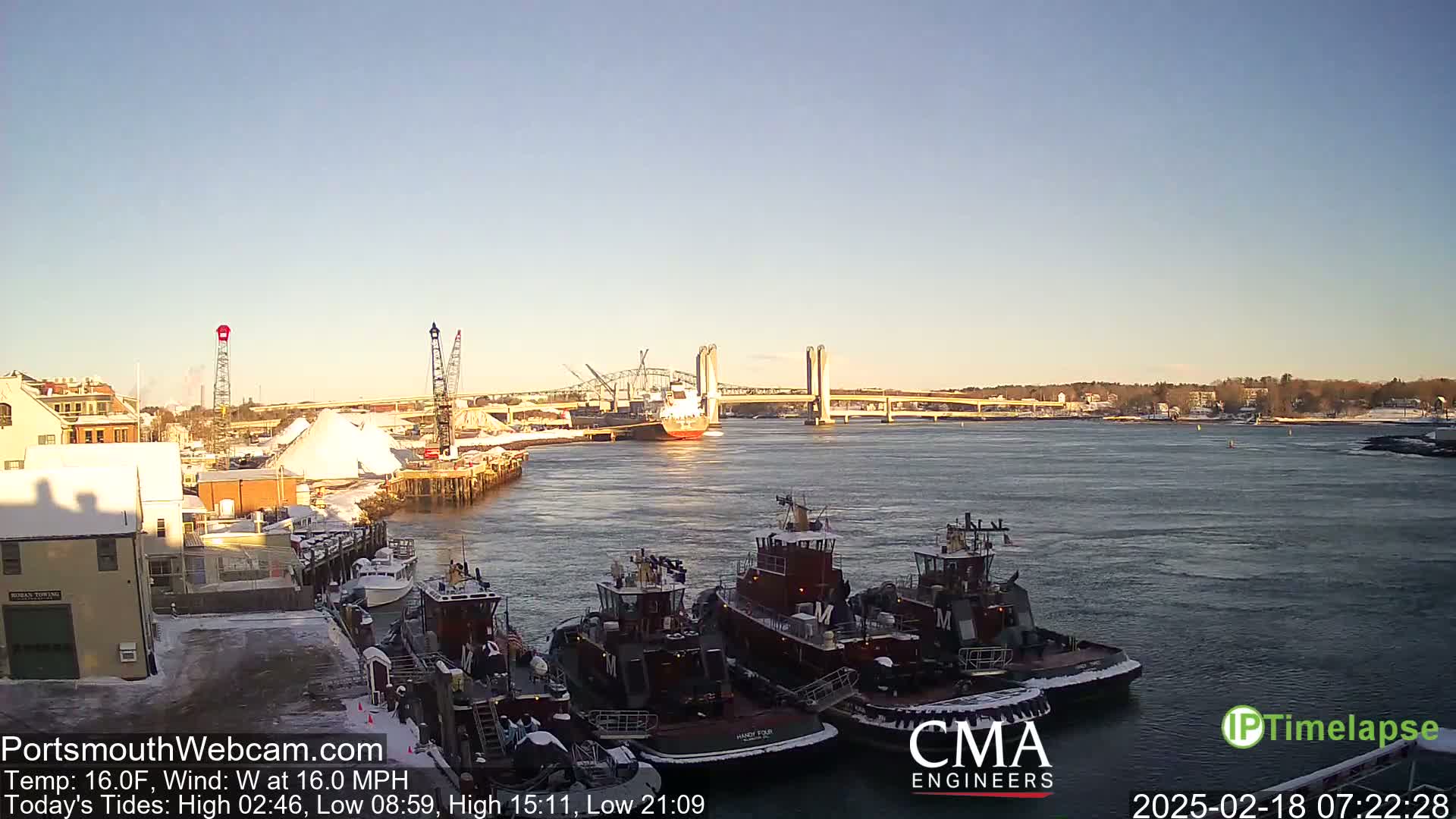 A harbor scene on a clear, cold day with several tugboats moored near a snow-covered dock, a large ship in the distance passing under a bridge.