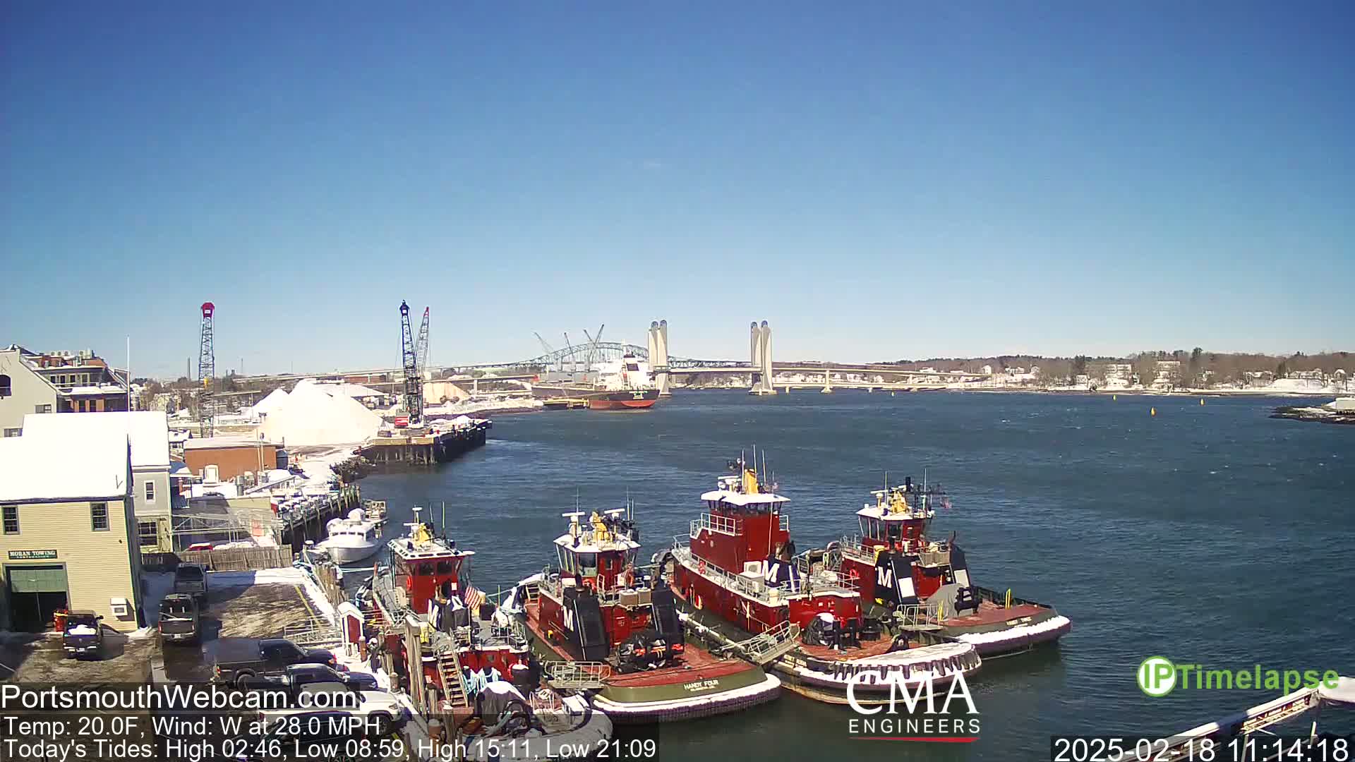 Several tugboats are moored at a dock next to a large pile of salt, with a bridge and calm, clear water in the background under a bright blue sky.