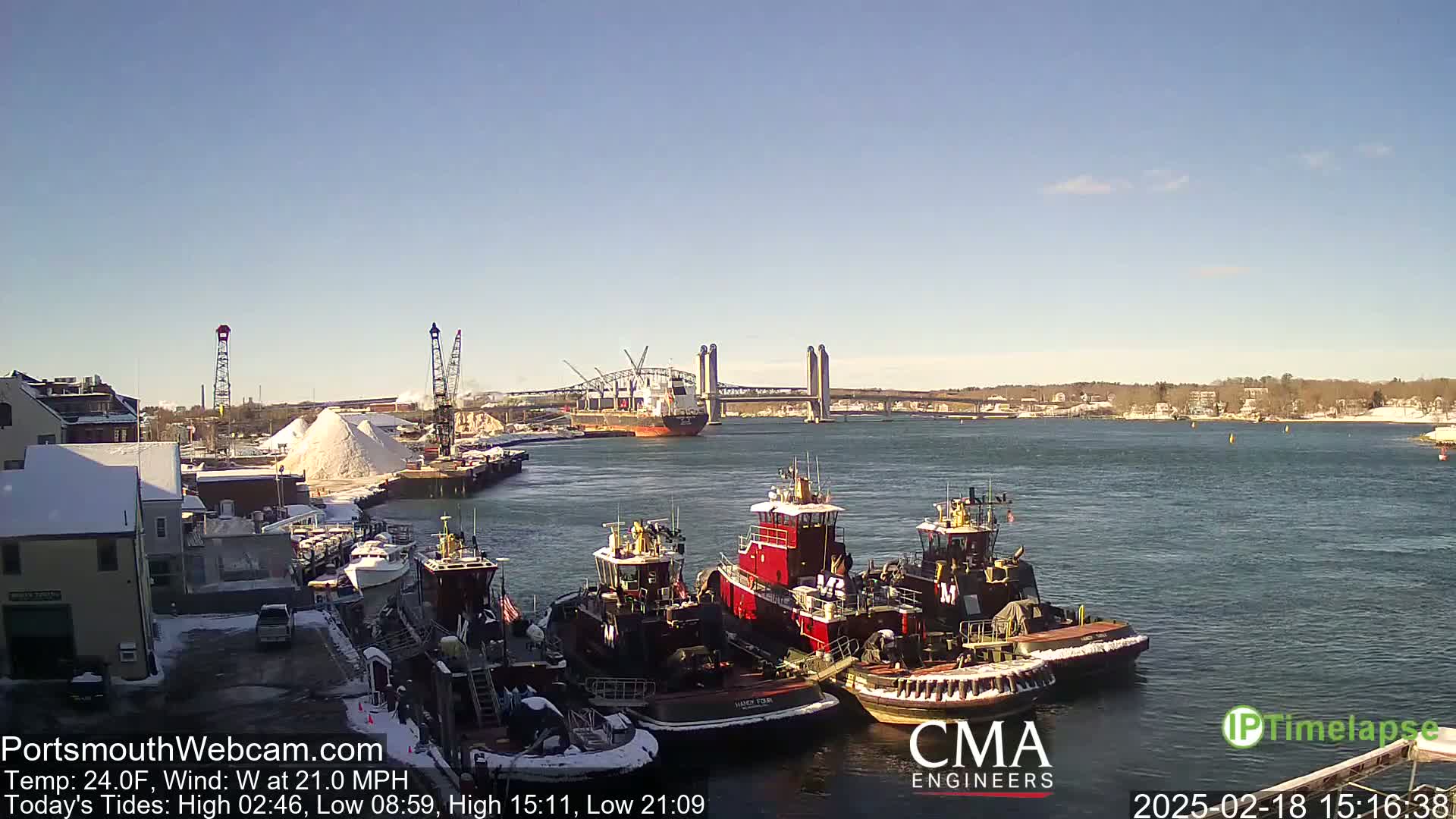 A clear, cold day reveals several tugboats docked near a snow-covered waterfront, with a large cargo ship and a bridge visible in the distance across a body of water.