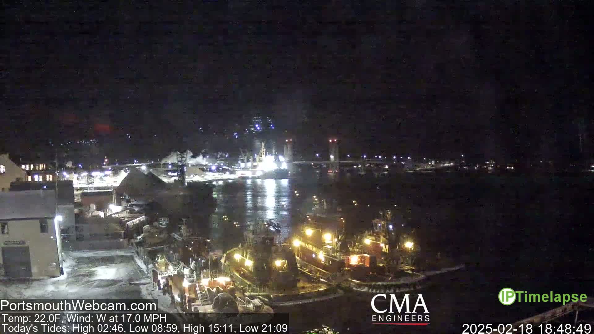 Several tugboats are moored near a lit-up waterfront at night, under a dark sky with a west wind.