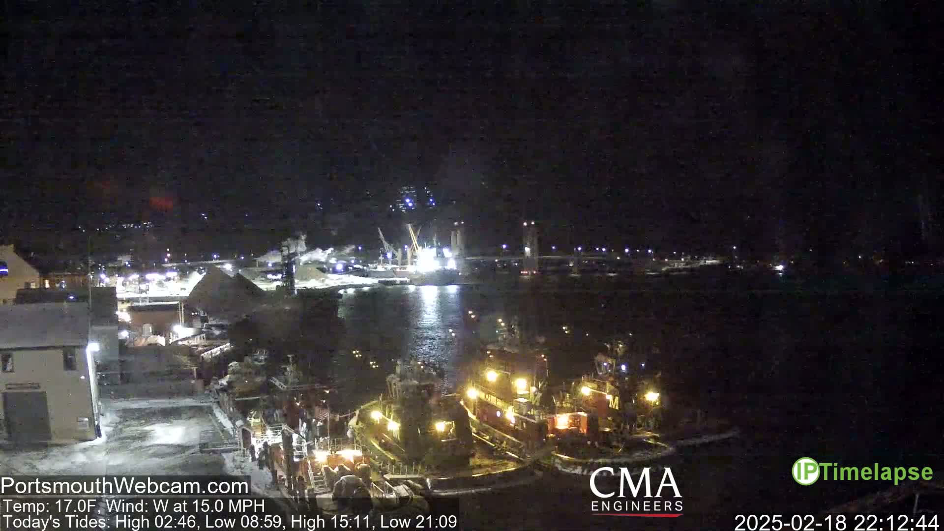 At night, several tugboats are docked near a waterfront industrial area under a dark sky with a west wind.