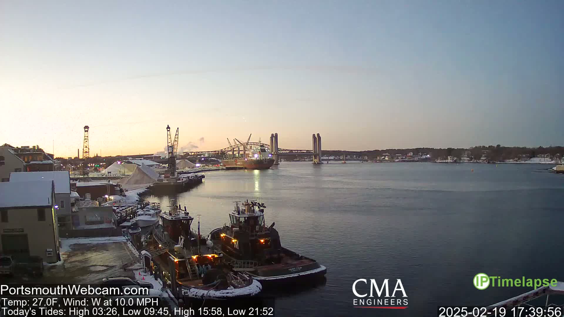 A harbor scene at dusk with a partly cloudy sky, snow on the ground, tugboats docked, a large cargo ship in the water, and a lift bridge in the distance.