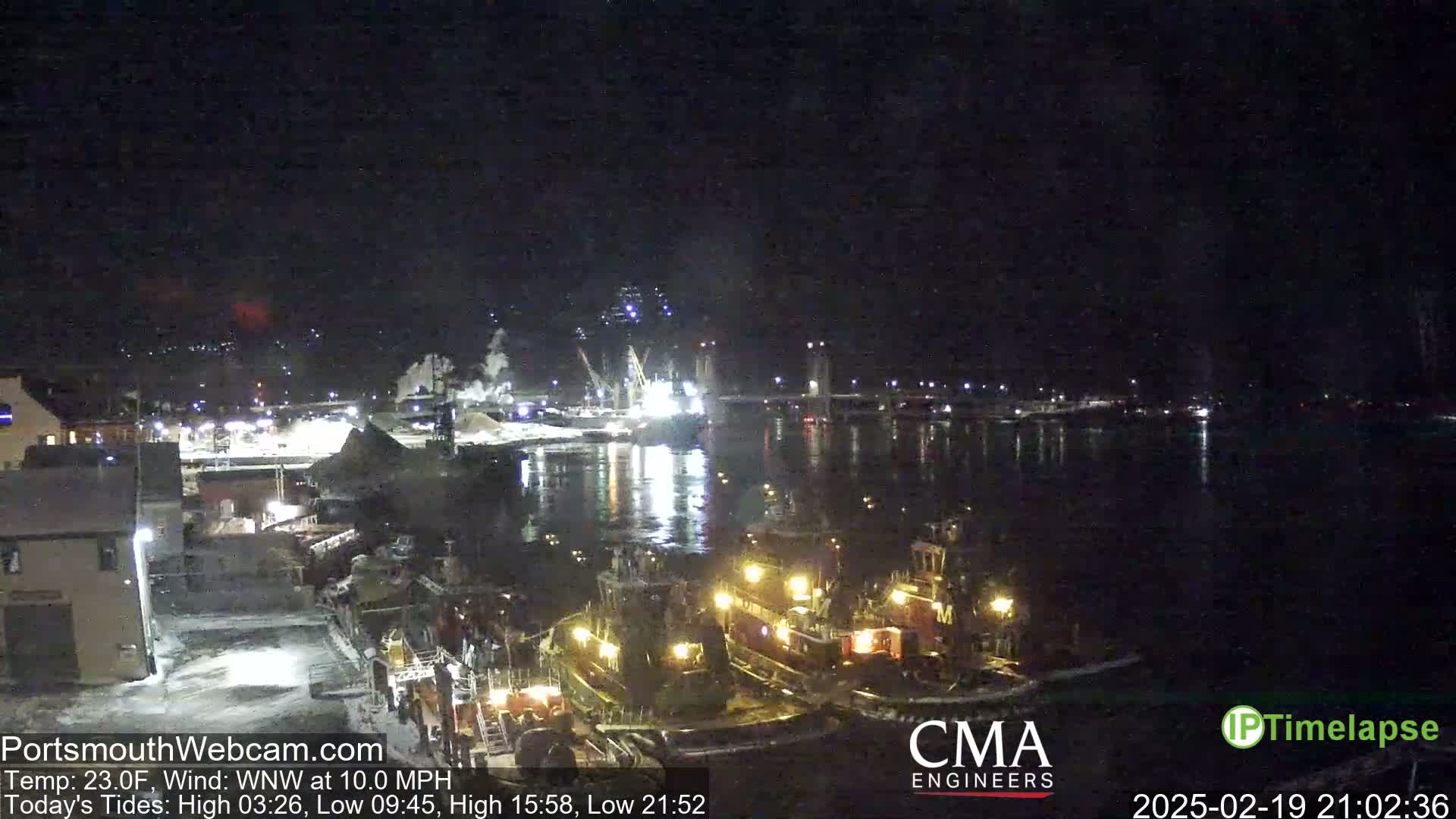 Several tugboats are moored at a dark waterfront at night, with a bridge and industrial area visible in the background under a clear, cold sky.