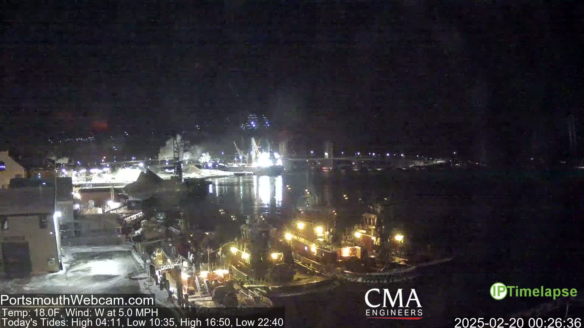 Several tugboats are moored at a nighttime harbor under a clear, dark sky, with industrial activity visible in the background.