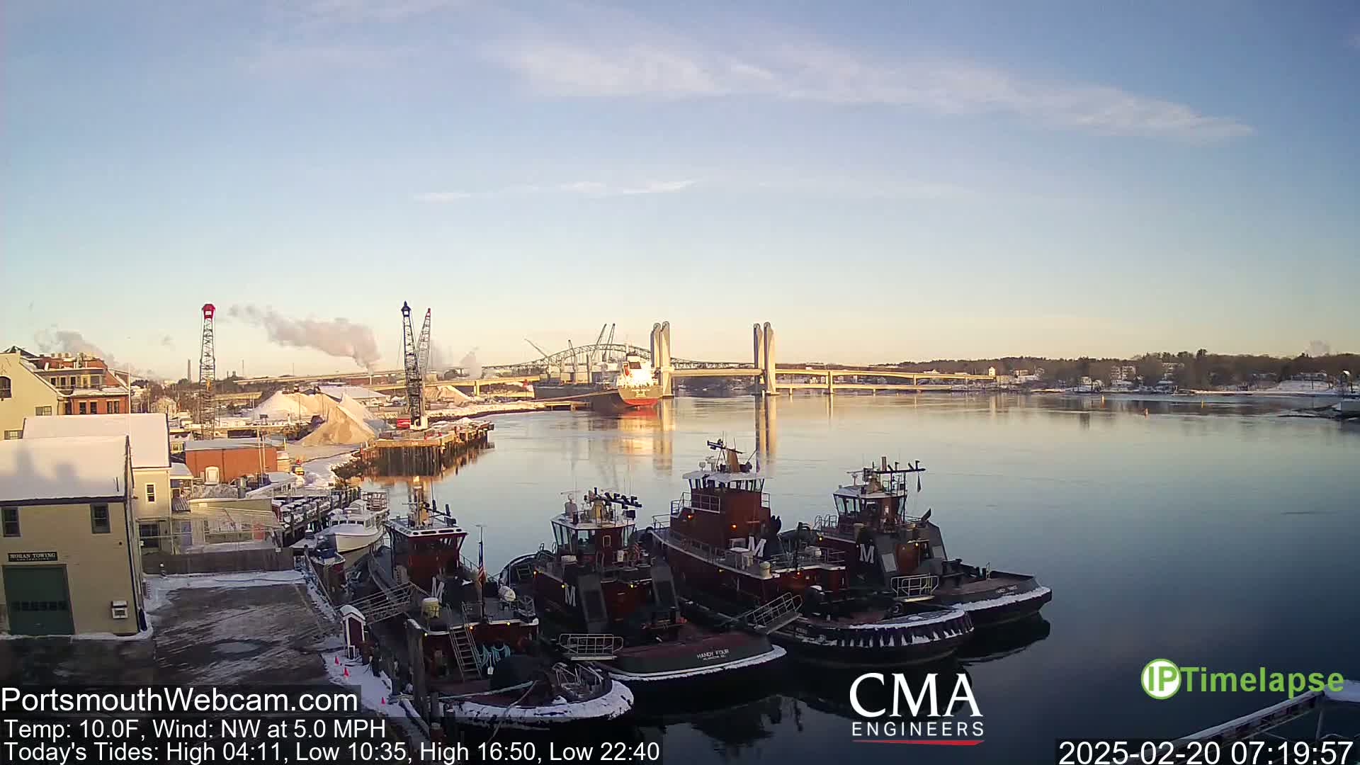 Several tugboats are moored at a dock in a calm, cold, snowy harbor, with a bridge and a large cargo ship visible in the background under a clear sky.