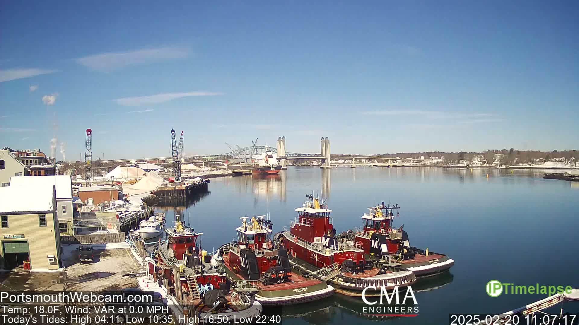 Several tugboats are docked at a waterfront location on a sunny, cold day, with a bridge and a larger vessel visible in the distance.