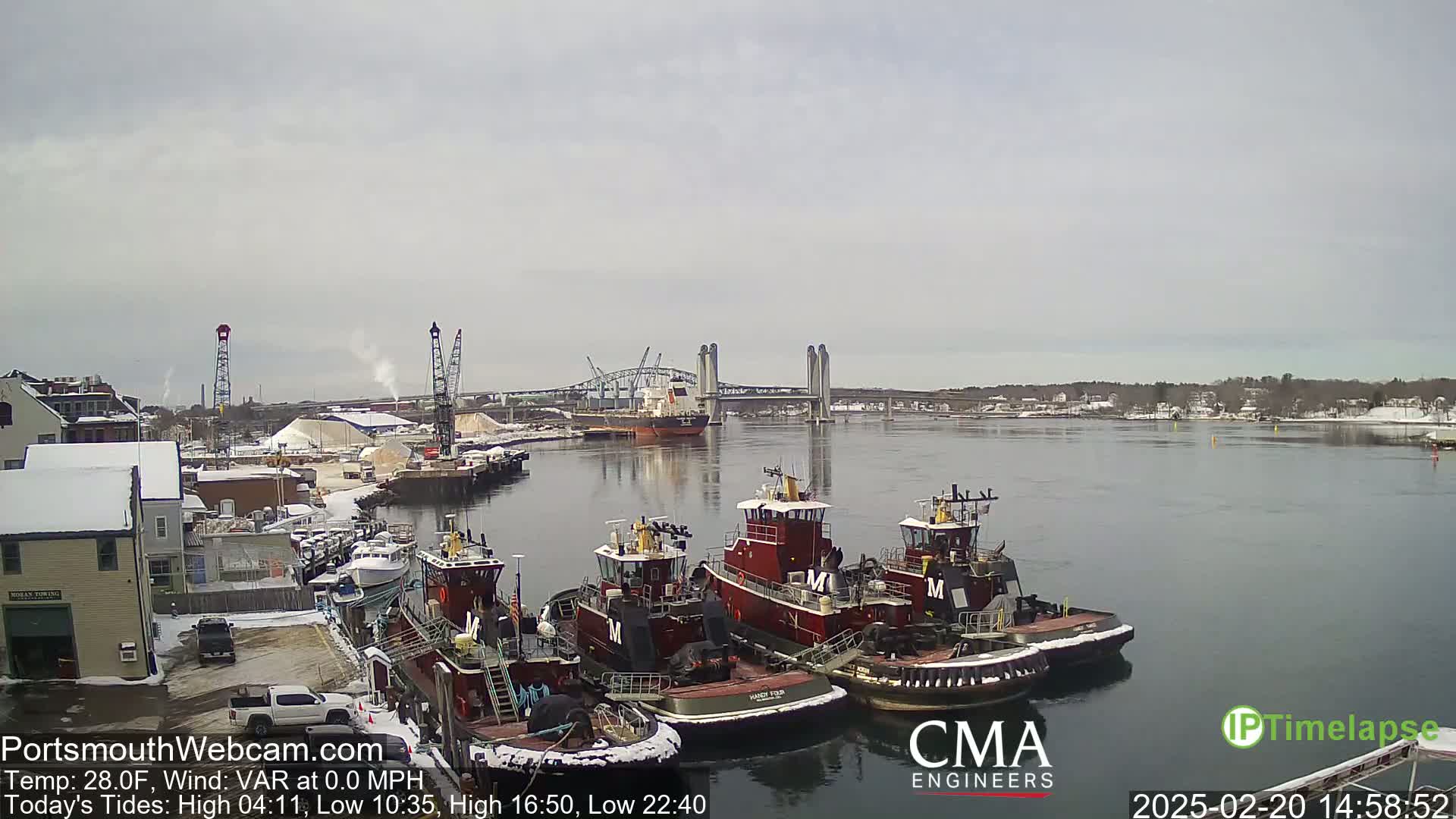 Several tugboats are moored at a dock next to a waterway, with a larger cargo ship and a bridge visible in the distance under a cloudy sky.