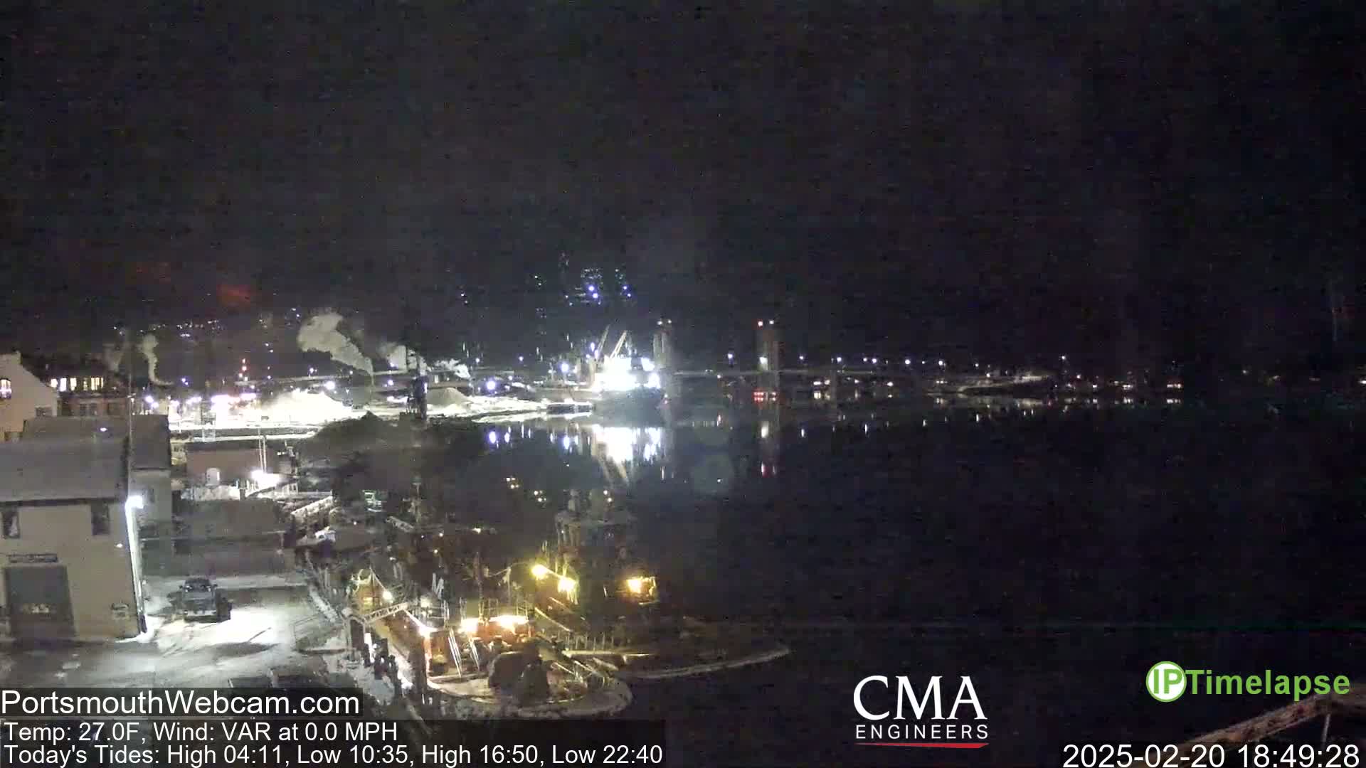 A nighttime view of a harbor with several boats docked, industrial buildings along the shore, and a bridge in the distance under a clear, dark sky.