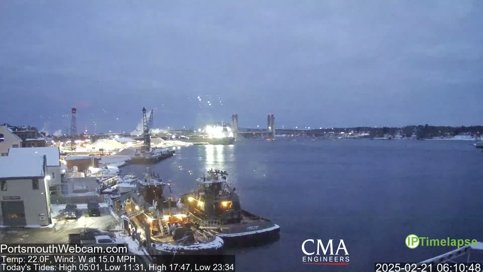 Two tugboats are docked near a snow-covered waterfront, with a larger vessel and a bridge visible in the distance under a twilight sky.