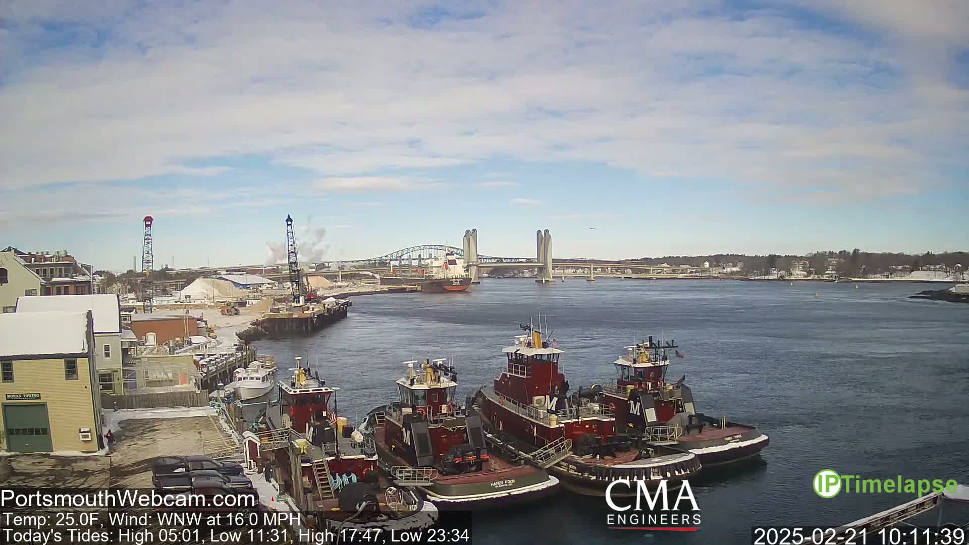 A partly cloudy day with a WNW wind at 16 mph shows several tugboats docked near a large cargo ship passing under a lift bridge in a snow-dusted harbor.