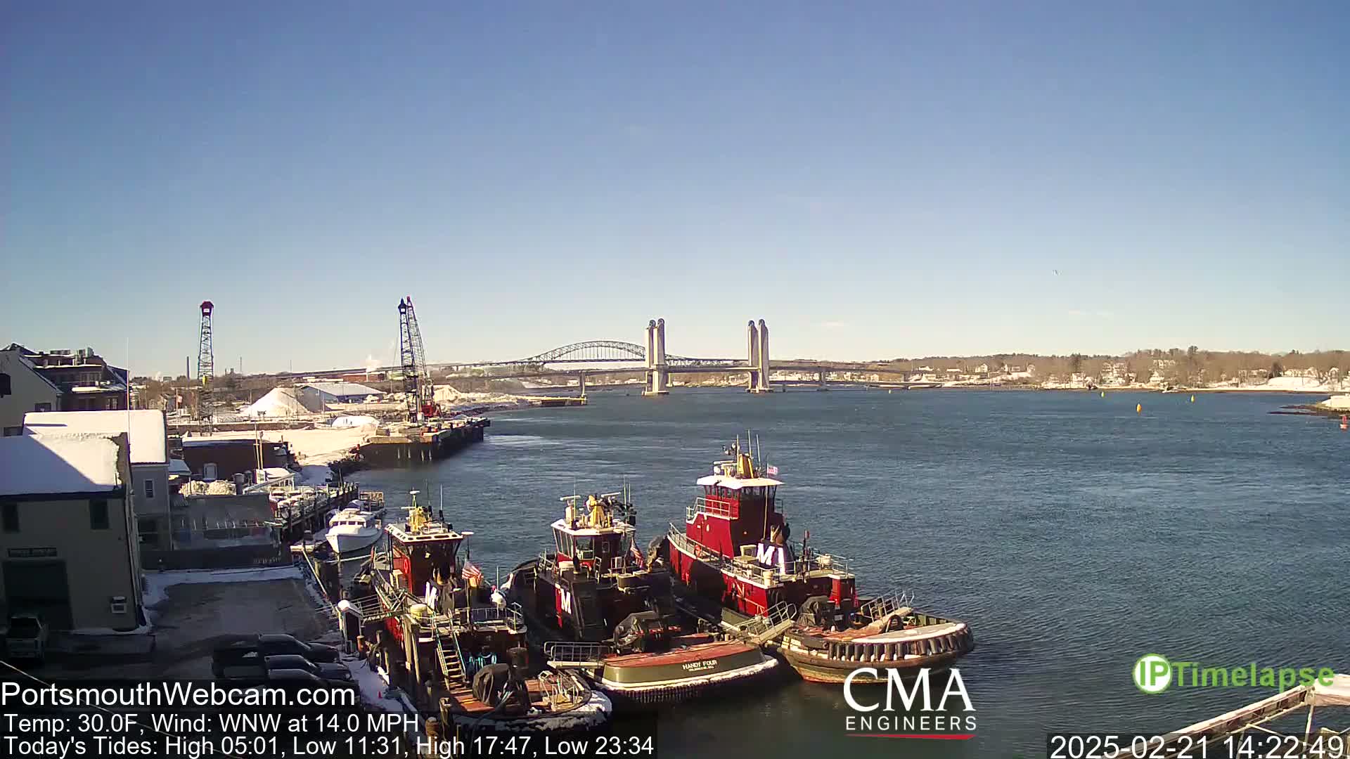 Several tugboats are docked near a snow-covered waterfront, with a bridge visible in the background under a clear blue sky.