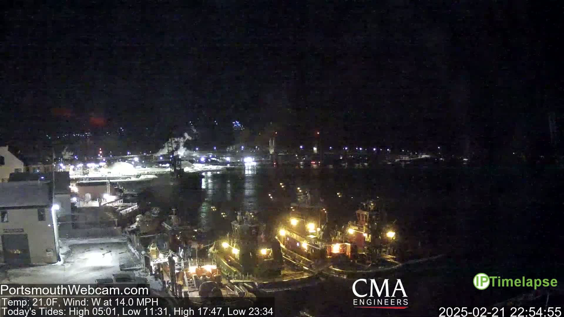 Several tugboats are moored at a dock at night in a harbor, under a dark sky with a west wind.