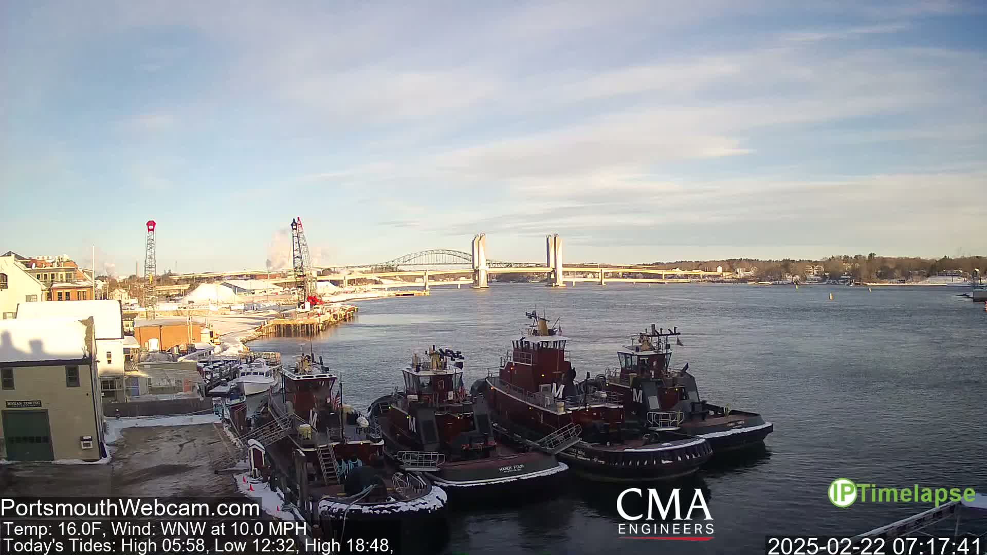 A harbor scene shows several tugboats docked near a waterfront, with a lift bridge in the distance under a partly cloudy sky.