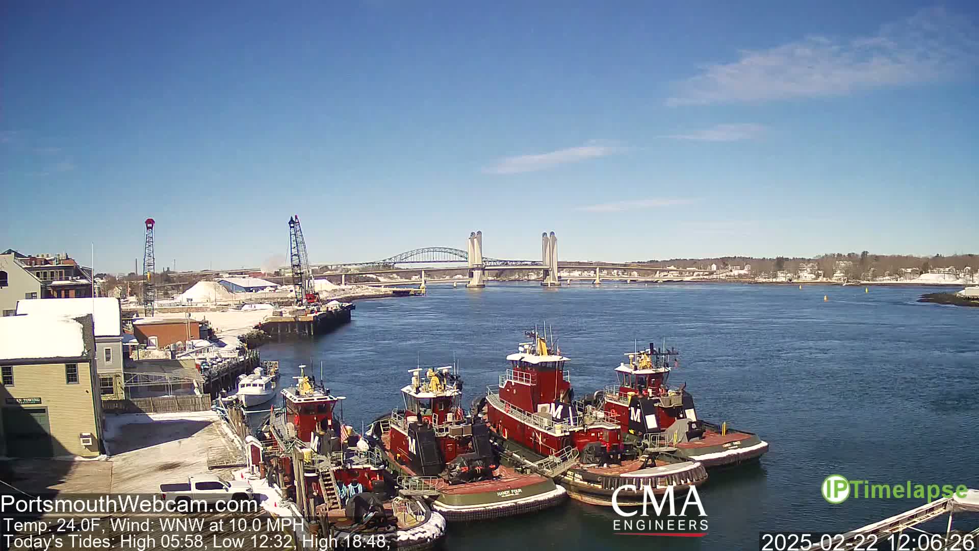 Several tugboats are moored at a dock near a bridge on a calm, sunny day.