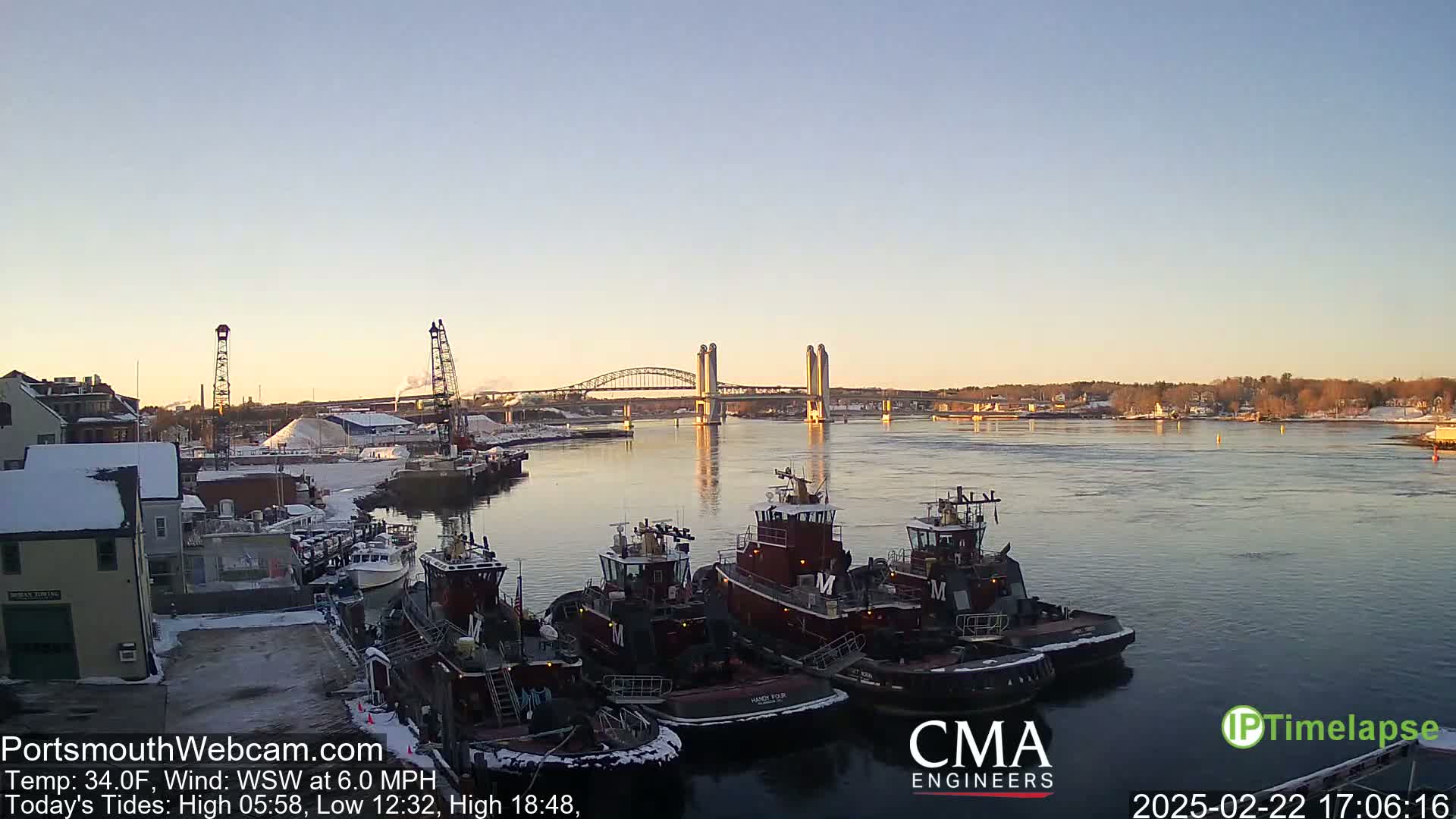 Several tugboats are moored at a snow-dusted waterfront, with a double-leaf bascule bridge in the background under a clear, twilight sky.