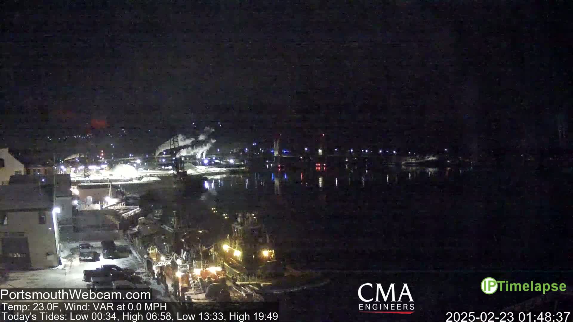 A nighttime view of a harbor with several boats and industrial buildings along the waterfront under a clear, dark sky.