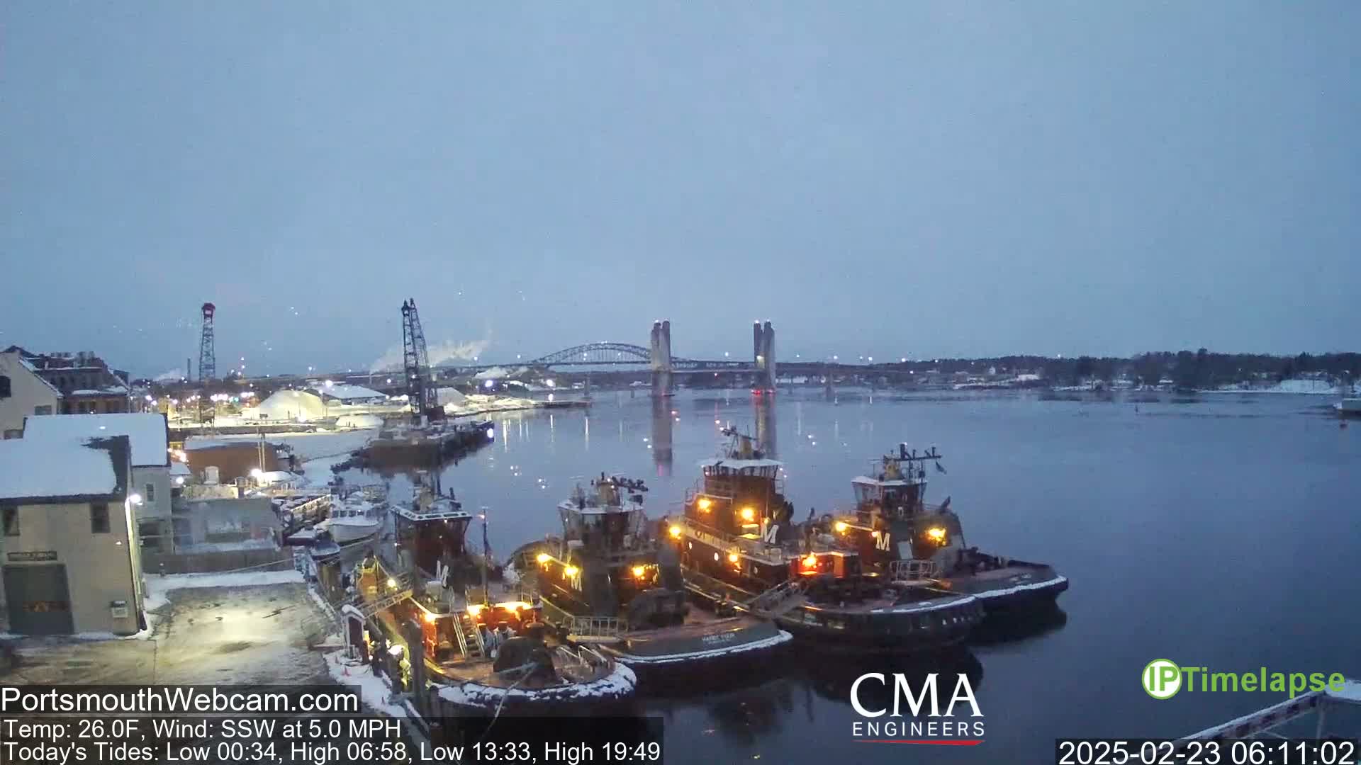 Three tugboats are moored at a snow-covered dock at twilight, with a bridge visible in the background across a calm river.