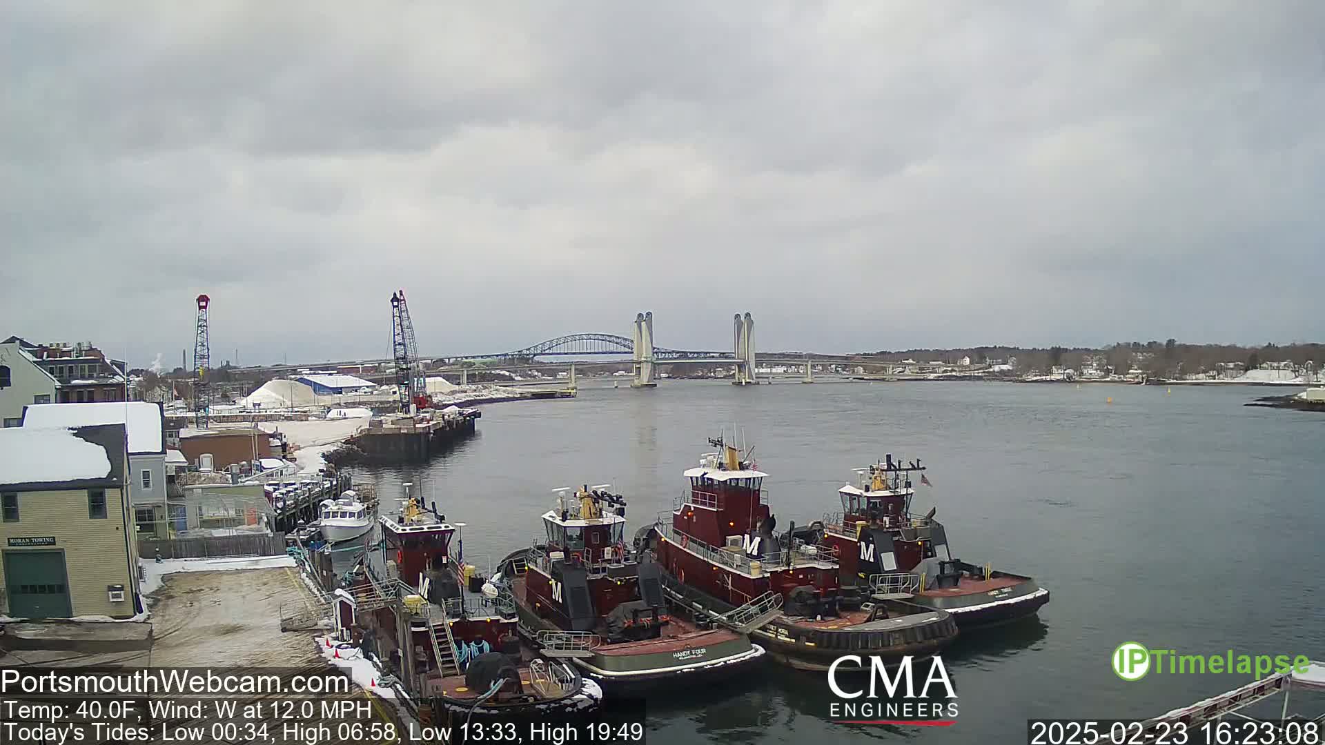Several tugboats are moored at a dock near a bridge on a cloudy day with a west wind.