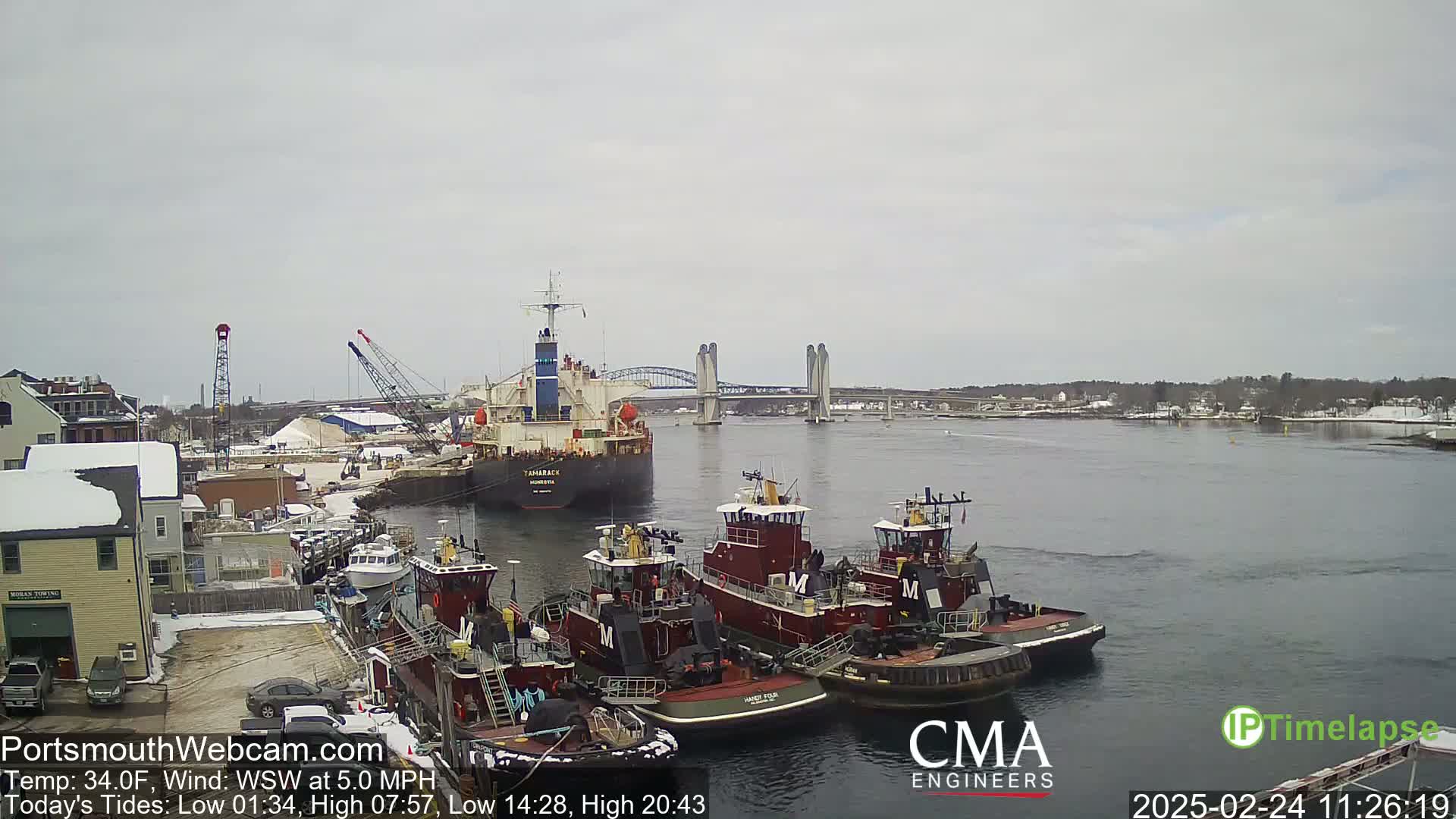 Several tugboats are moored at a dock next to a larger cargo ship, under a cloudy sky, with a lift bridge in the distance.