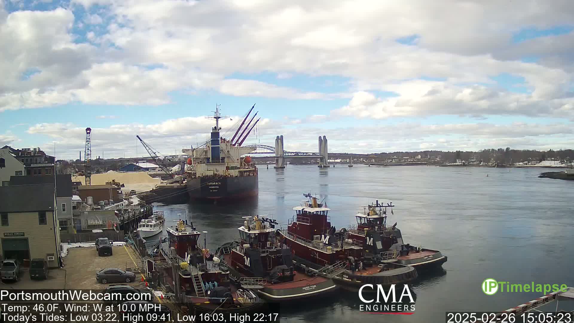 A large cargo ship and several tugboats are docked at a waterfront, under a partly cloudy sky, with a lift bridge in the background.