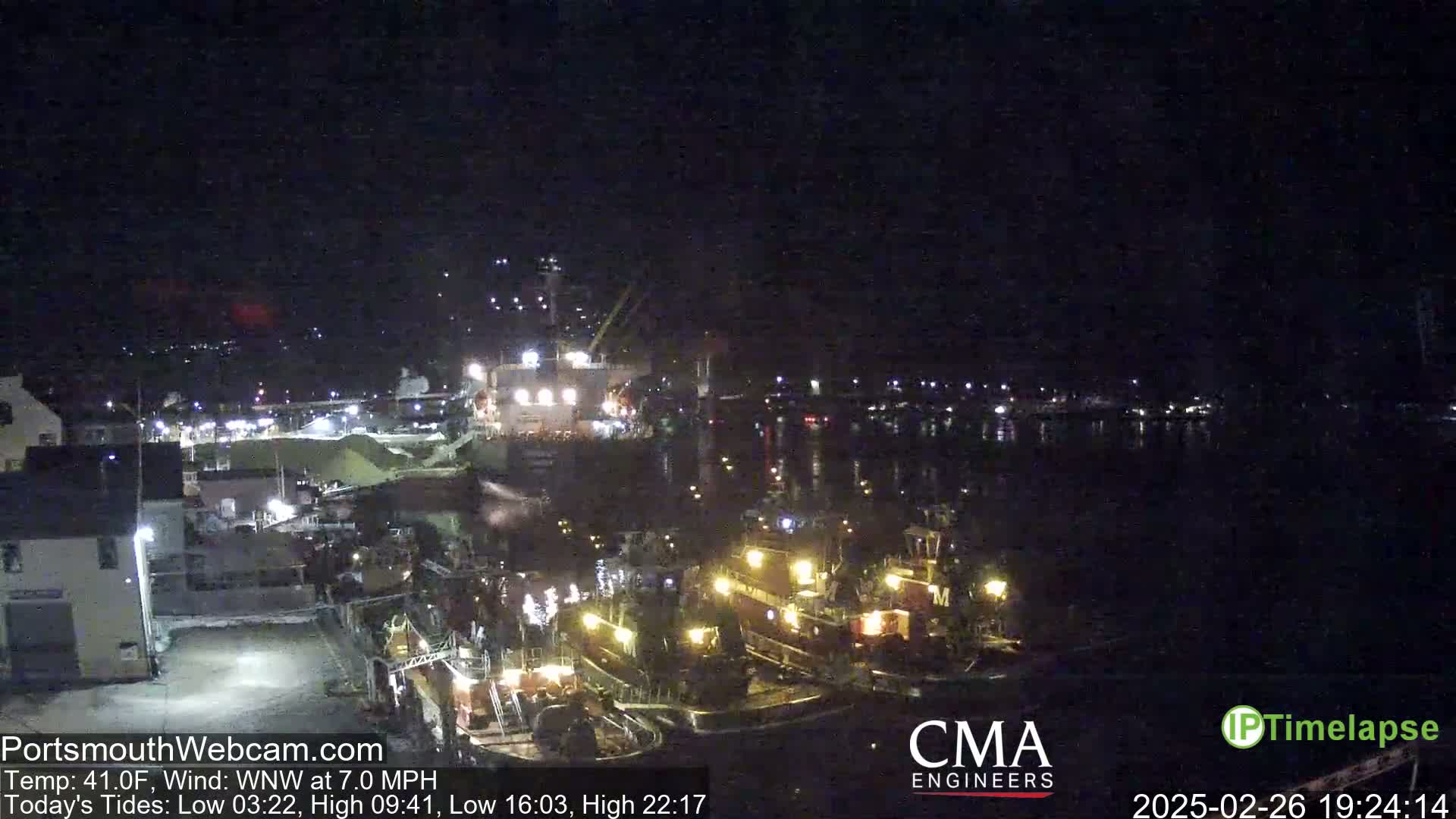 A nighttime harbor scene shows several docked boats illuminated by lights, under a dark sky with a wind of 7.0 mph.