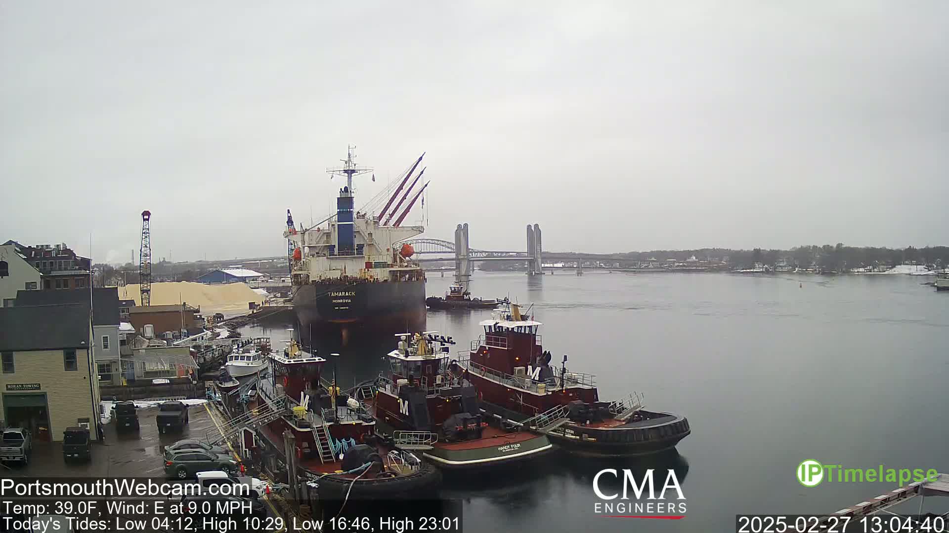 A large cargo ship is docked near several tugboats, with a bridge in the background, under an overcast sky.