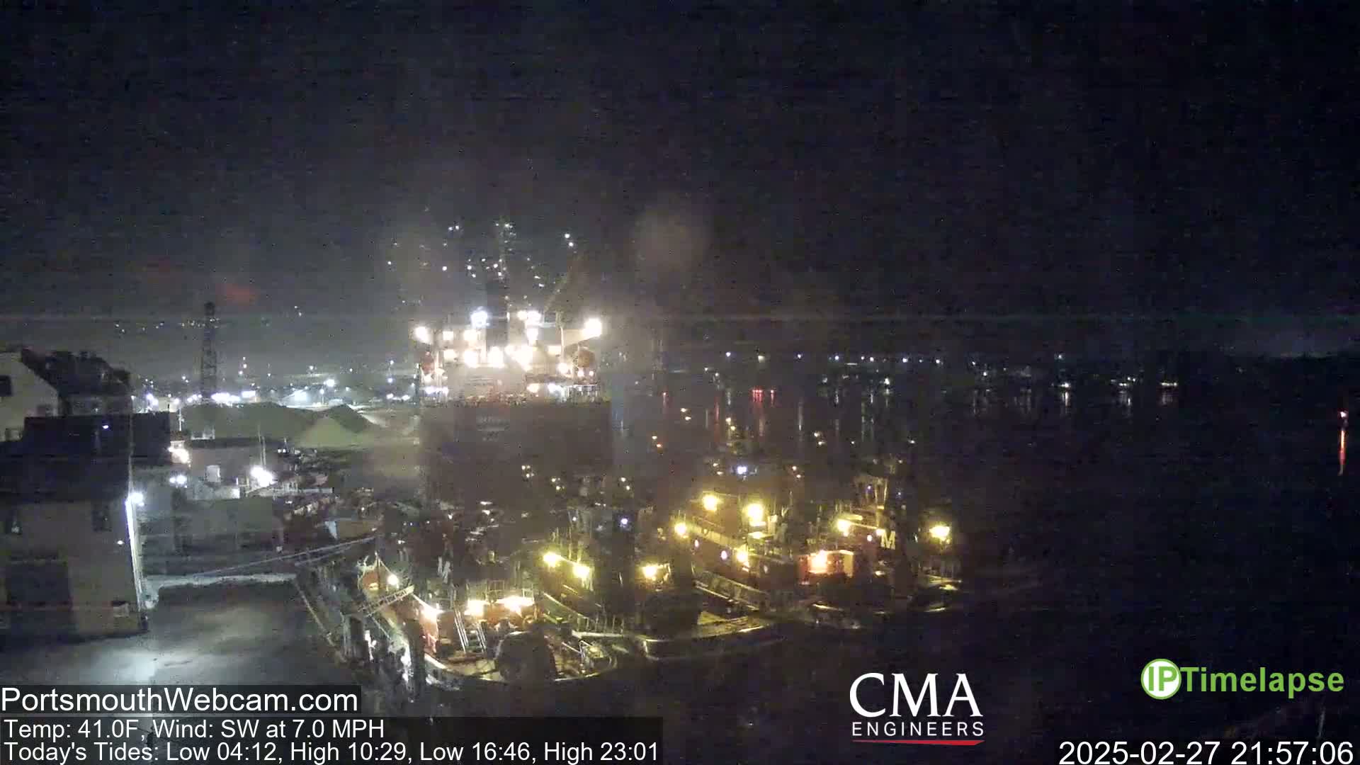 A large cargo ship is being assisted by several tugboats in a harbor at night under a dark sky.
