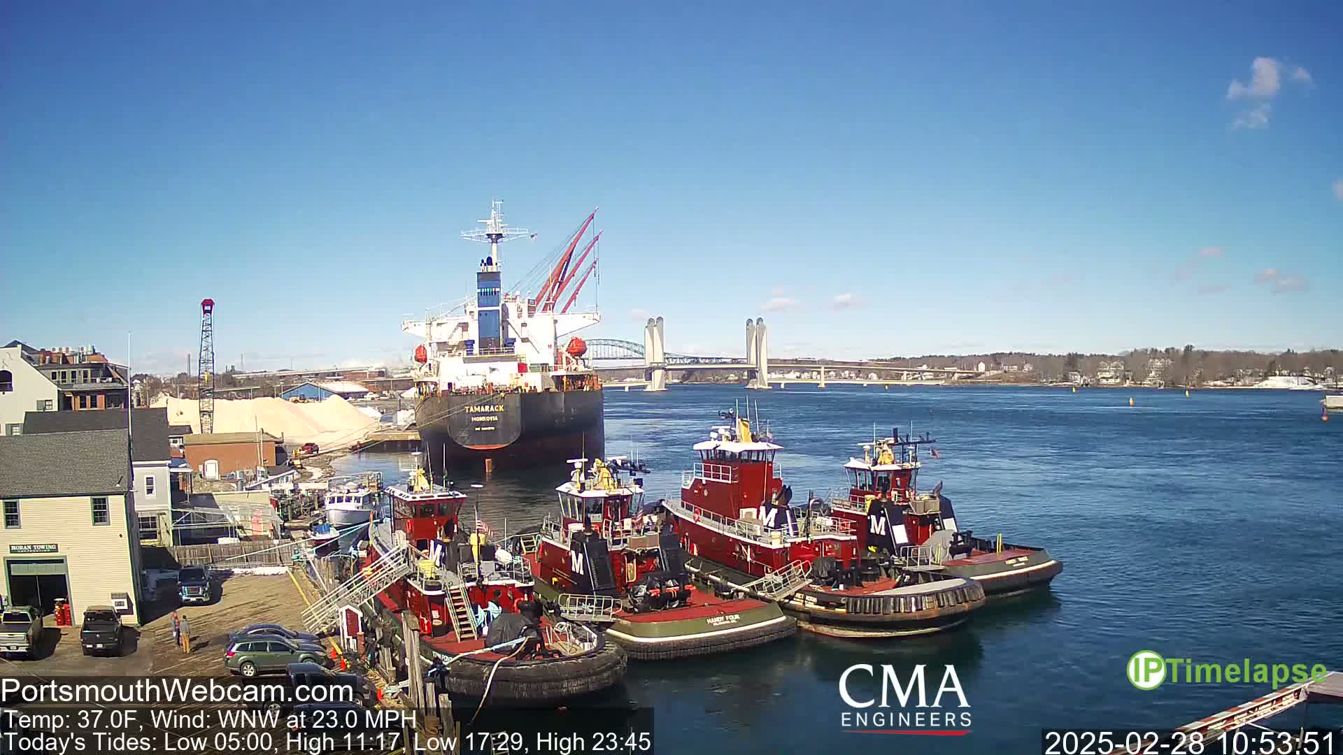 A large cargo ship is moored near several tugboats in a harbor under a clear blue sky, with a bridge visible in the distance and a 23 mph wind.