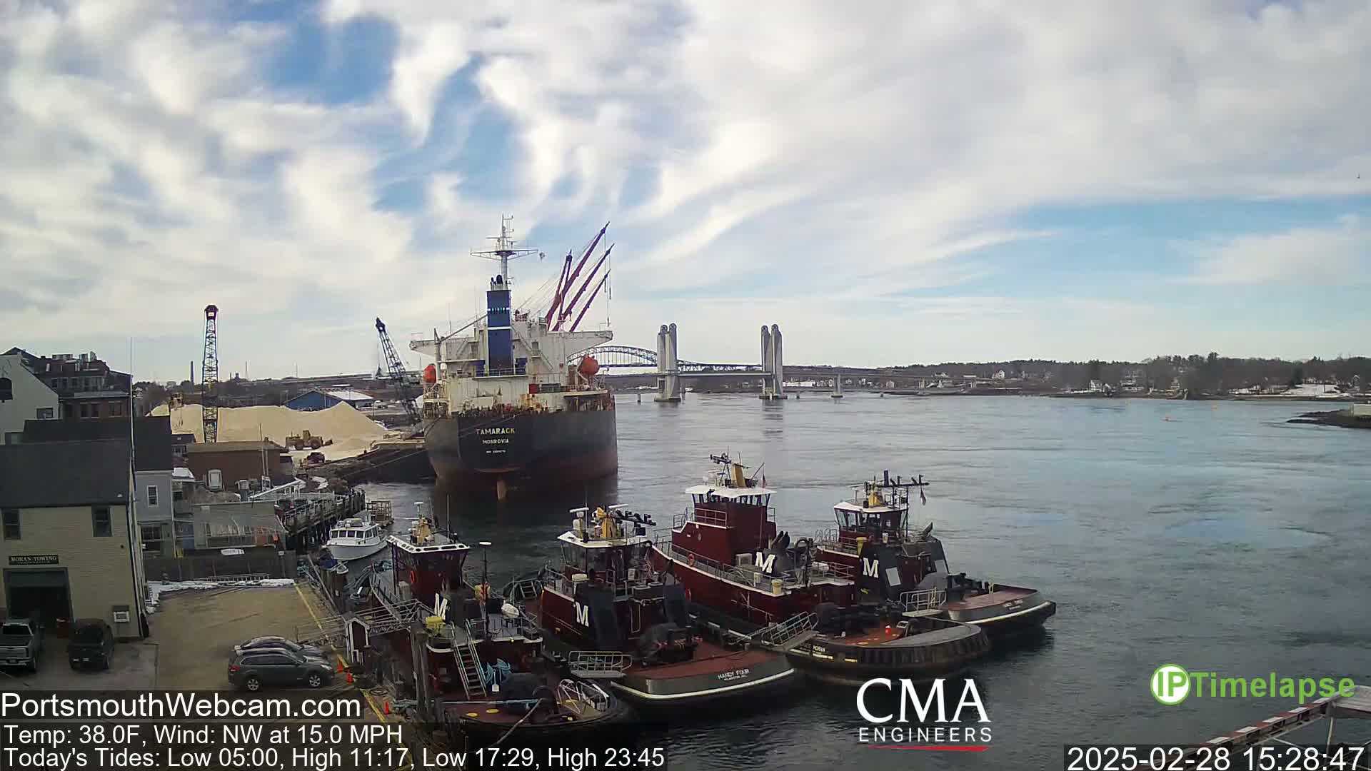 A large cargo ship is docked near several tugboats in a harbor under a partly cloudy sky, with a lift bridge visible in the distance.