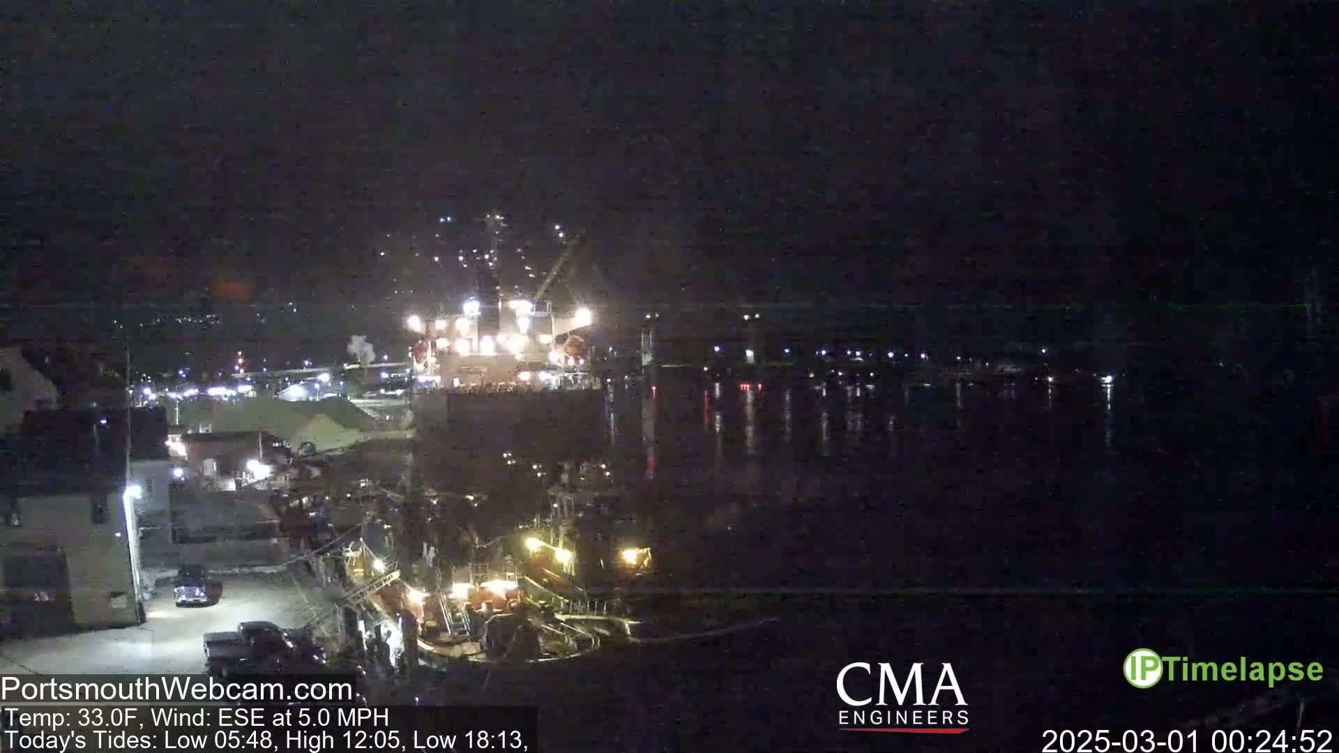A nighttime harbor scene shows a large ship with lights on, docked next to smaller vessels, under a dark, clear sky.