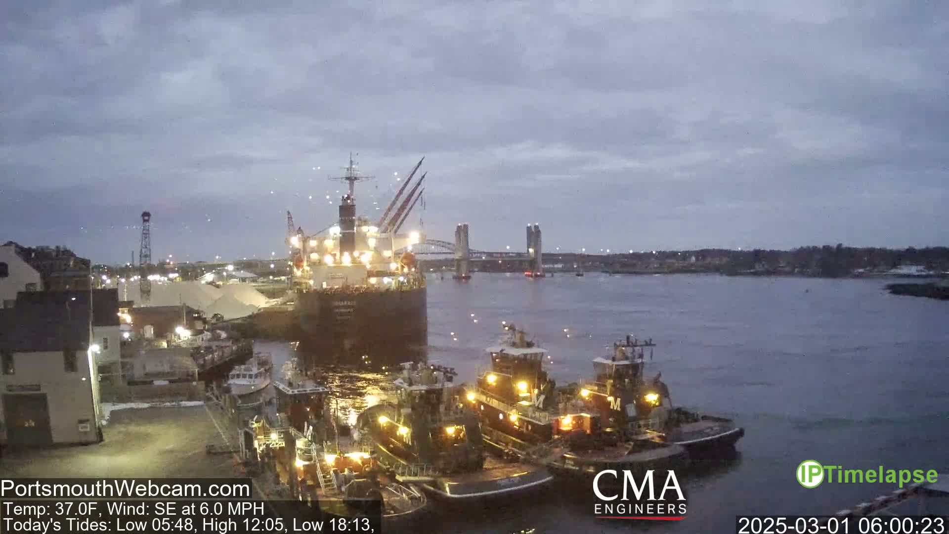 A large cargo ship is docked next to several tugboats in a harbor under a cloudy, twilight sky with a light southeast wind.