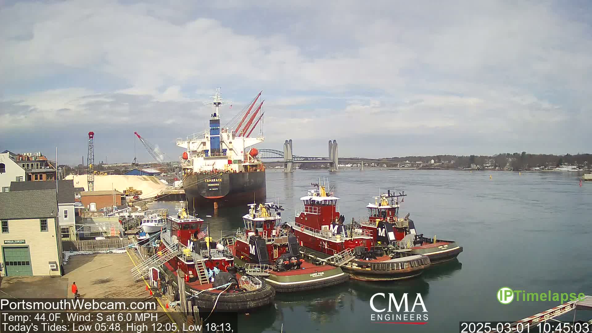 A large cargo ship is docked near several tugboats in a harbor under a partly cloudy sky.