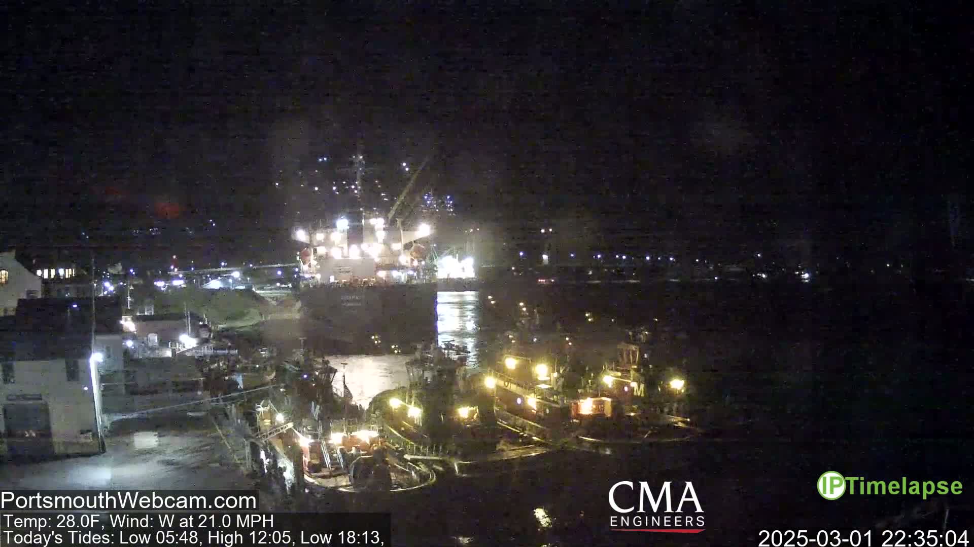 A large ship is being assisted by several tugboats in a harbor at night, under a dark sky with a 28°F temperature and a 21 mph west wind.
