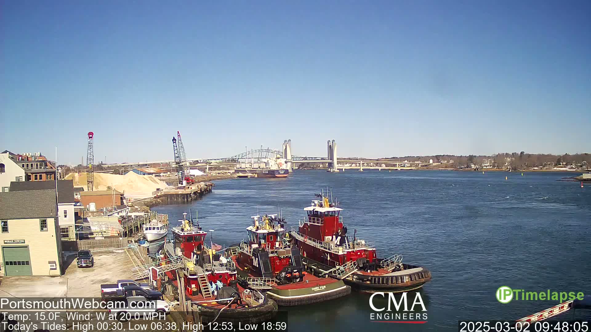Several tugboats are moored at a dock next to a body of water under a clear blue sky, with a large bridge visible in the distance.