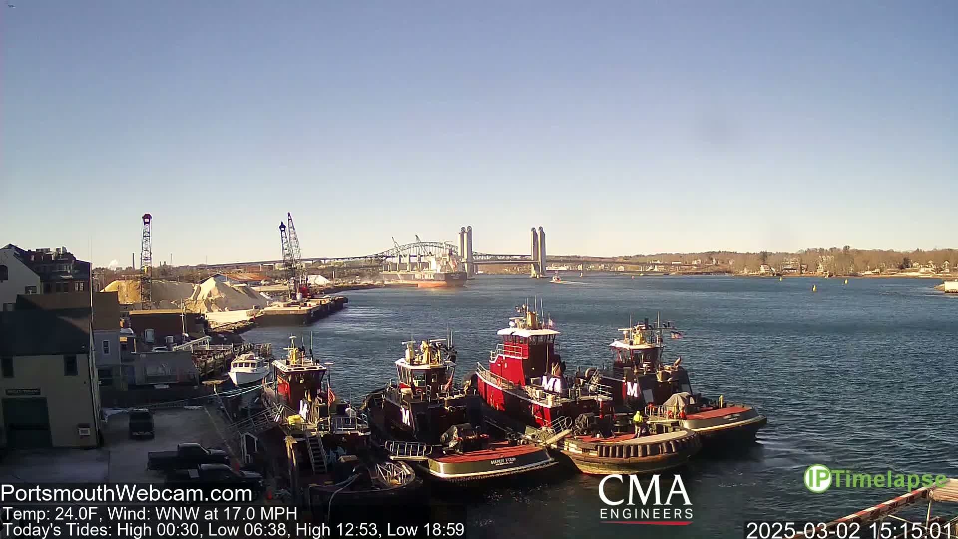 Several tugboats are moored at a dock near a large bridge, under a clear, sunny sky.