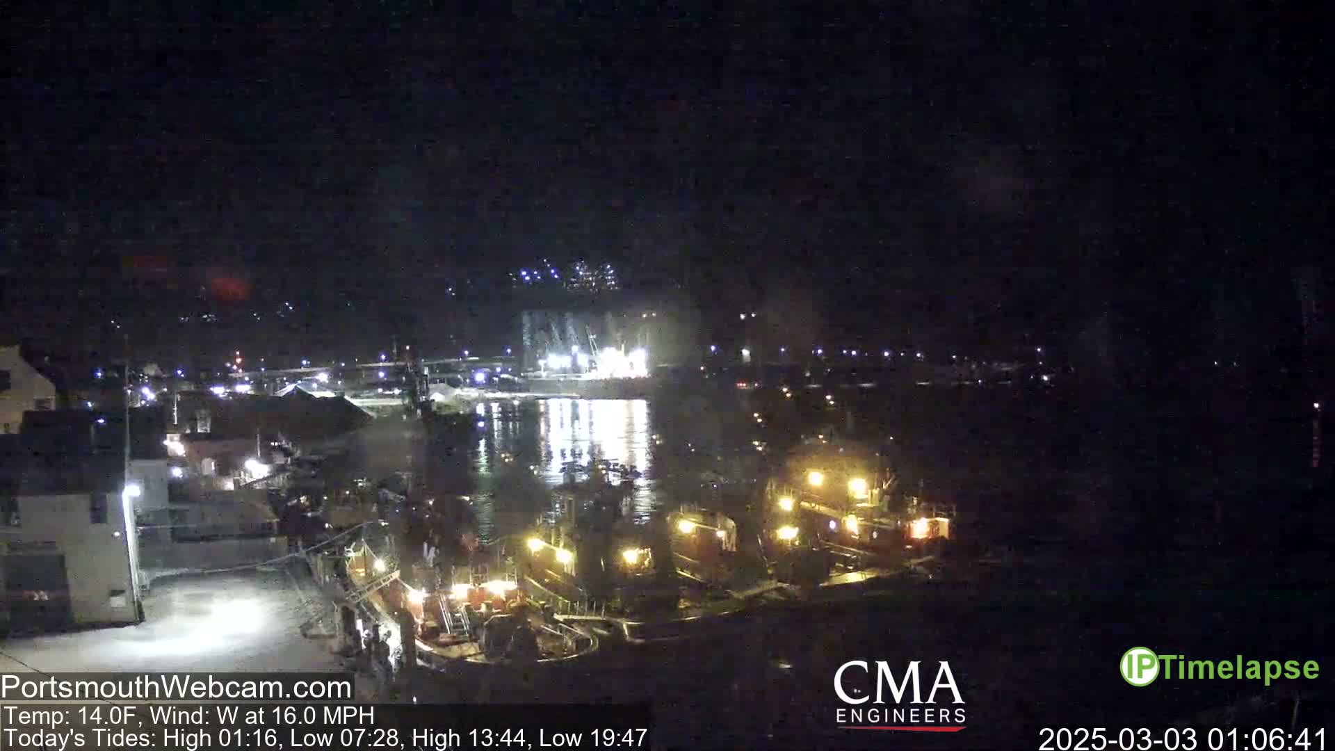 At night, under a dark sky with a west wind of 16 mph and a temperature of 14°F, several lit-up tugboats are moored in a harbor near a waterfront area with buildings.