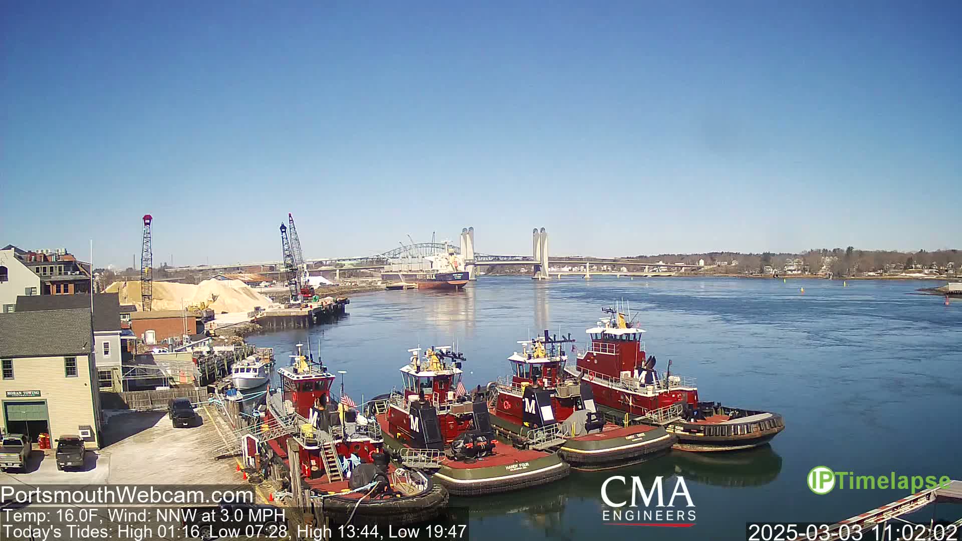 Several tugboats are moored at a dock beside a river under a clear blue sky, with a large bridge and a cargo ship visible in the distance.