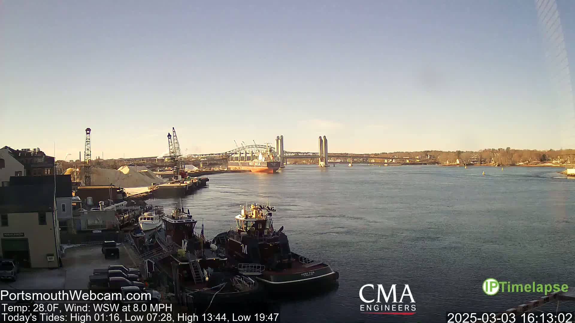 A large cargo ship is passing under a bridge on a calm, sunny day near a harbor with tugboats and industrial areas visible on the shore.