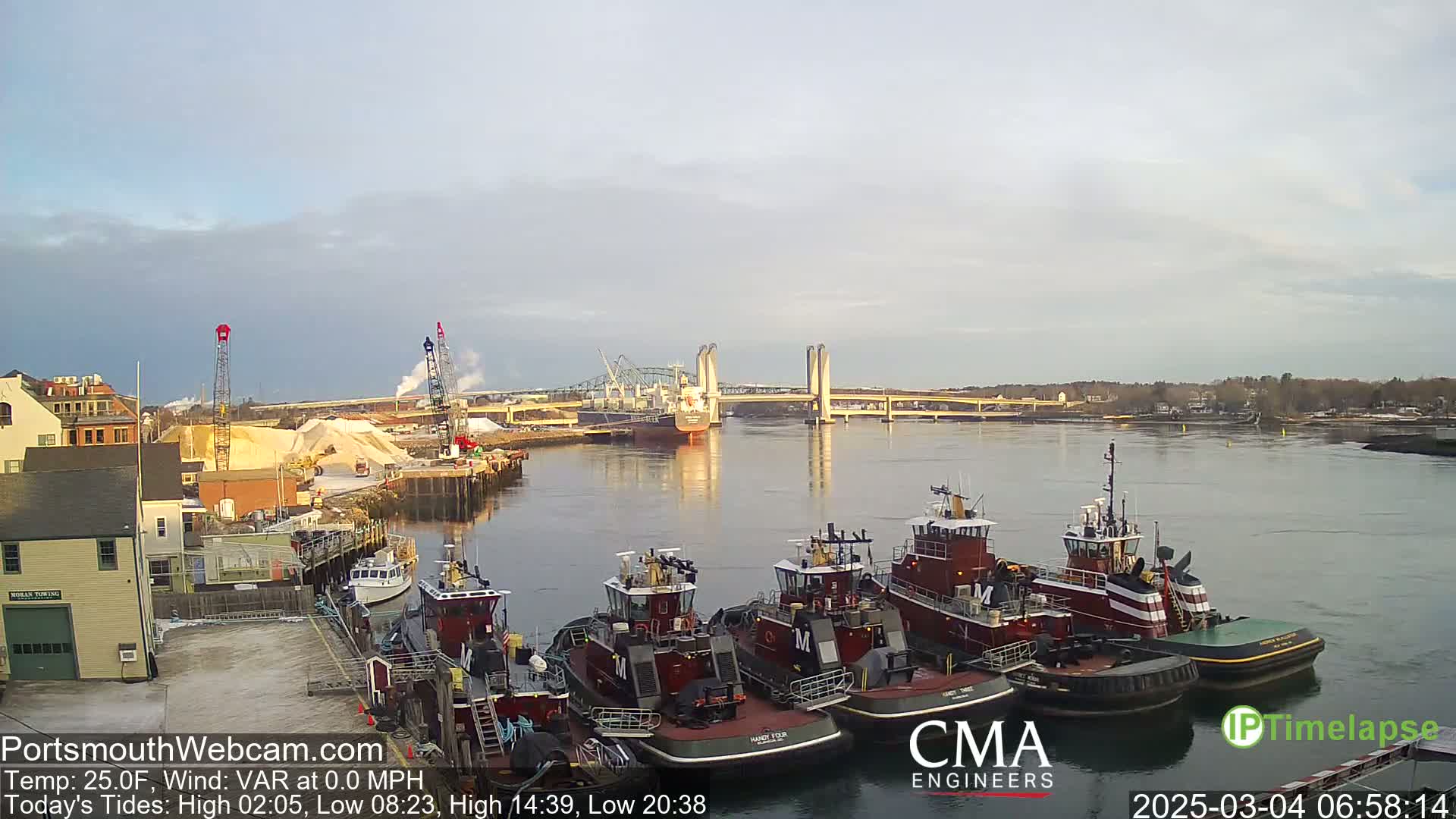 Several tugboats are moored along a waterfront, with a larger vessel and a lift bridge visible in the distance across a calm river under a partly cloudy sky.