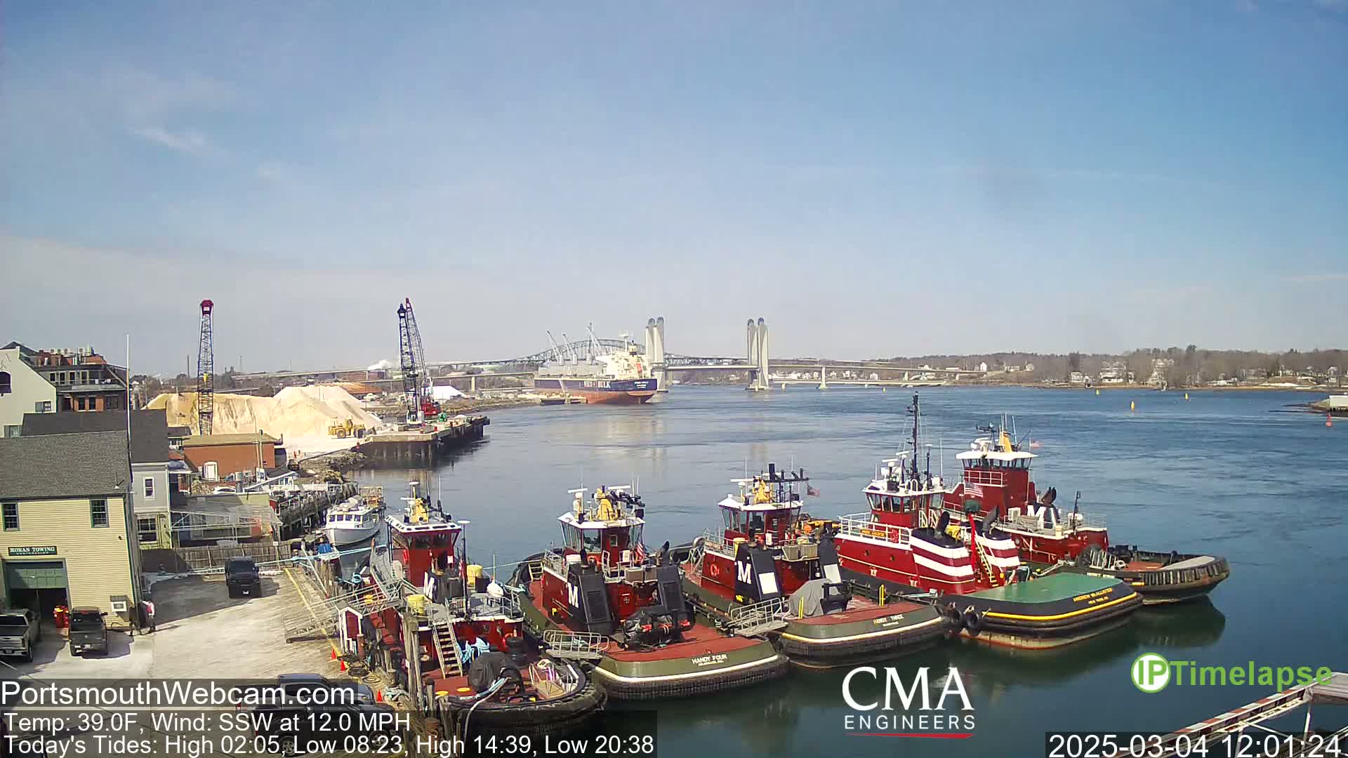 Several tugboats are moored at a dock next to a large pile of sand, with a large cargo ship and a bridge visible in the distance on a sunny day.