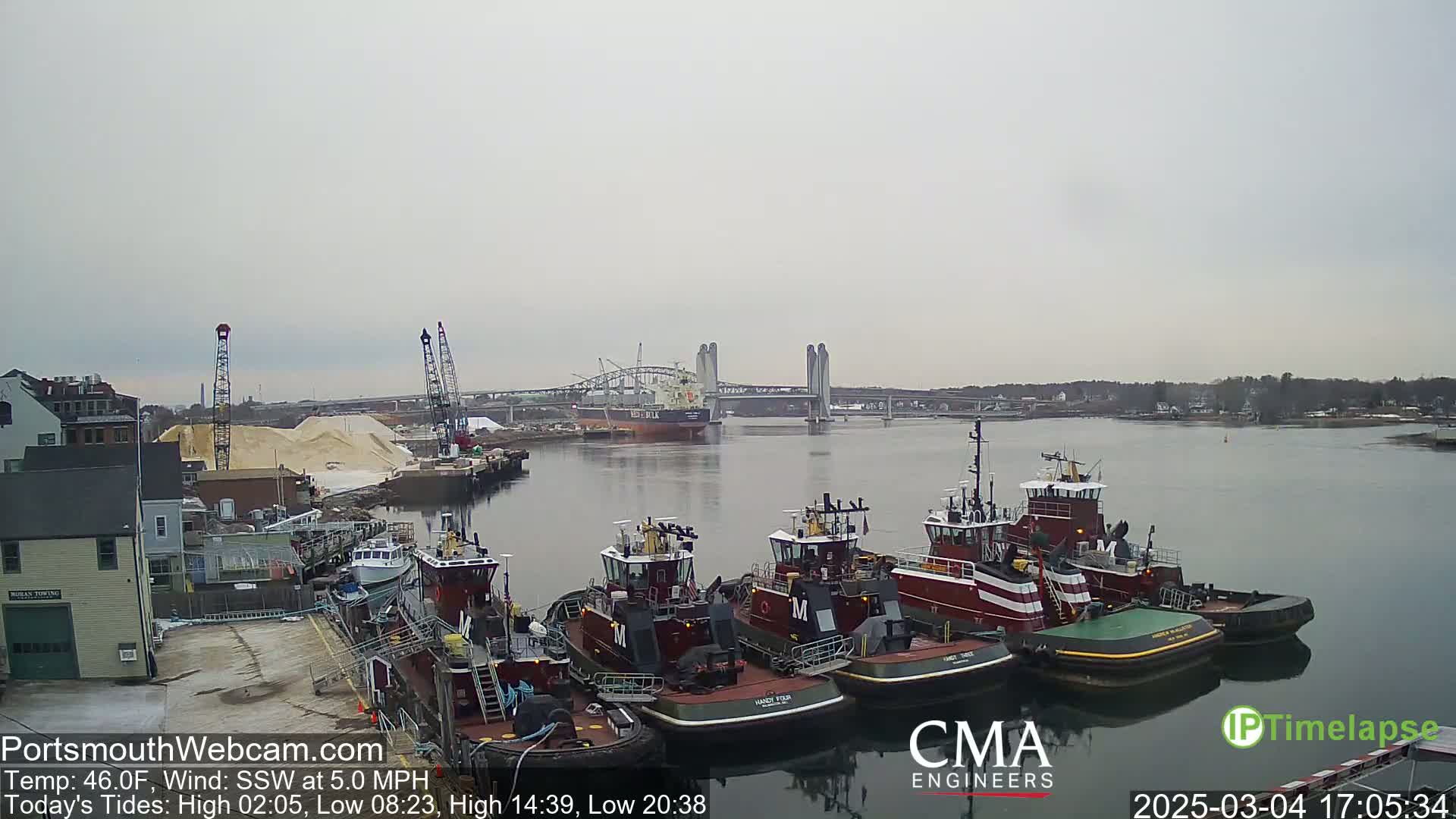 Several tugboats are docked at a waterfront industrial area on an overcast day, with a large ship and a bridge visible in the distance.